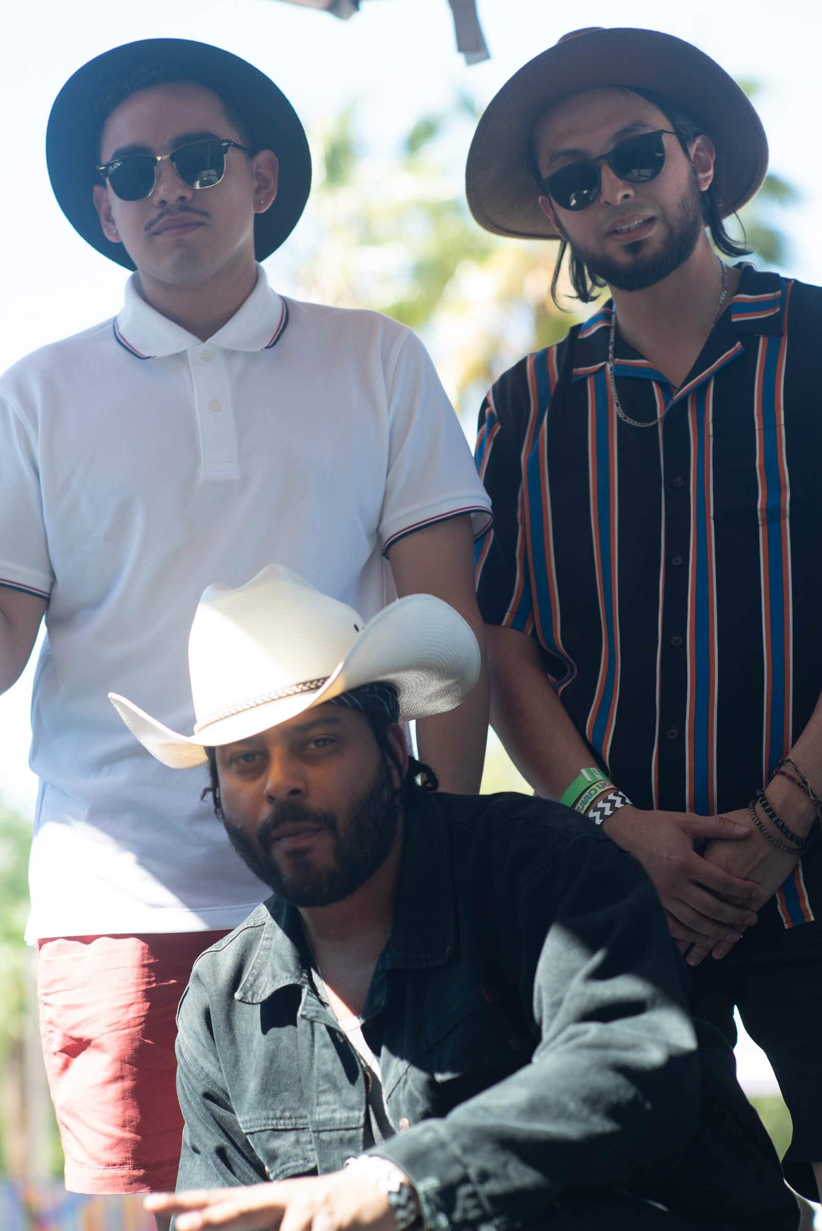 Cuatro hombres posando al aire libre en un día soleado. Uno lleva sombrero de vaquero y chaqueta negra, otro sombrero de ala ancha y camisa con rayas, y dos llevan sombrero negro y gafas de sol.