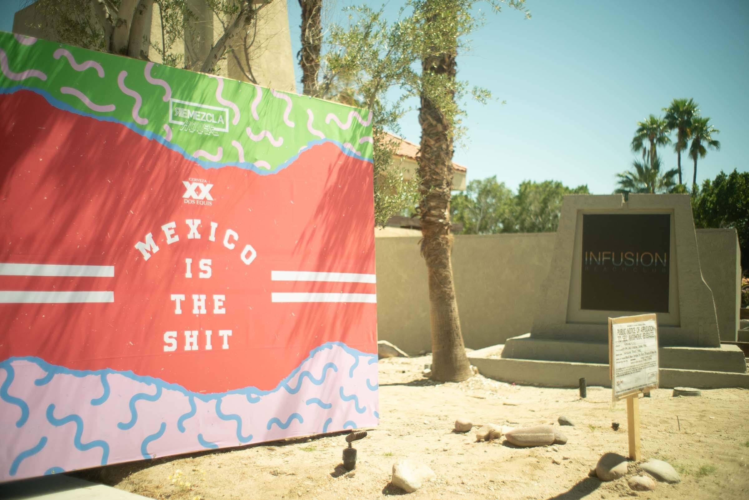 Una lona colorida con la frase "México is the shit" en letras blancas, decorada con líneas azules y verdes. Al fondo hay árboles y una estructura con una pantalla negra y un cartel informativo. El escenario parece ser al aire libre en un día soleado.
