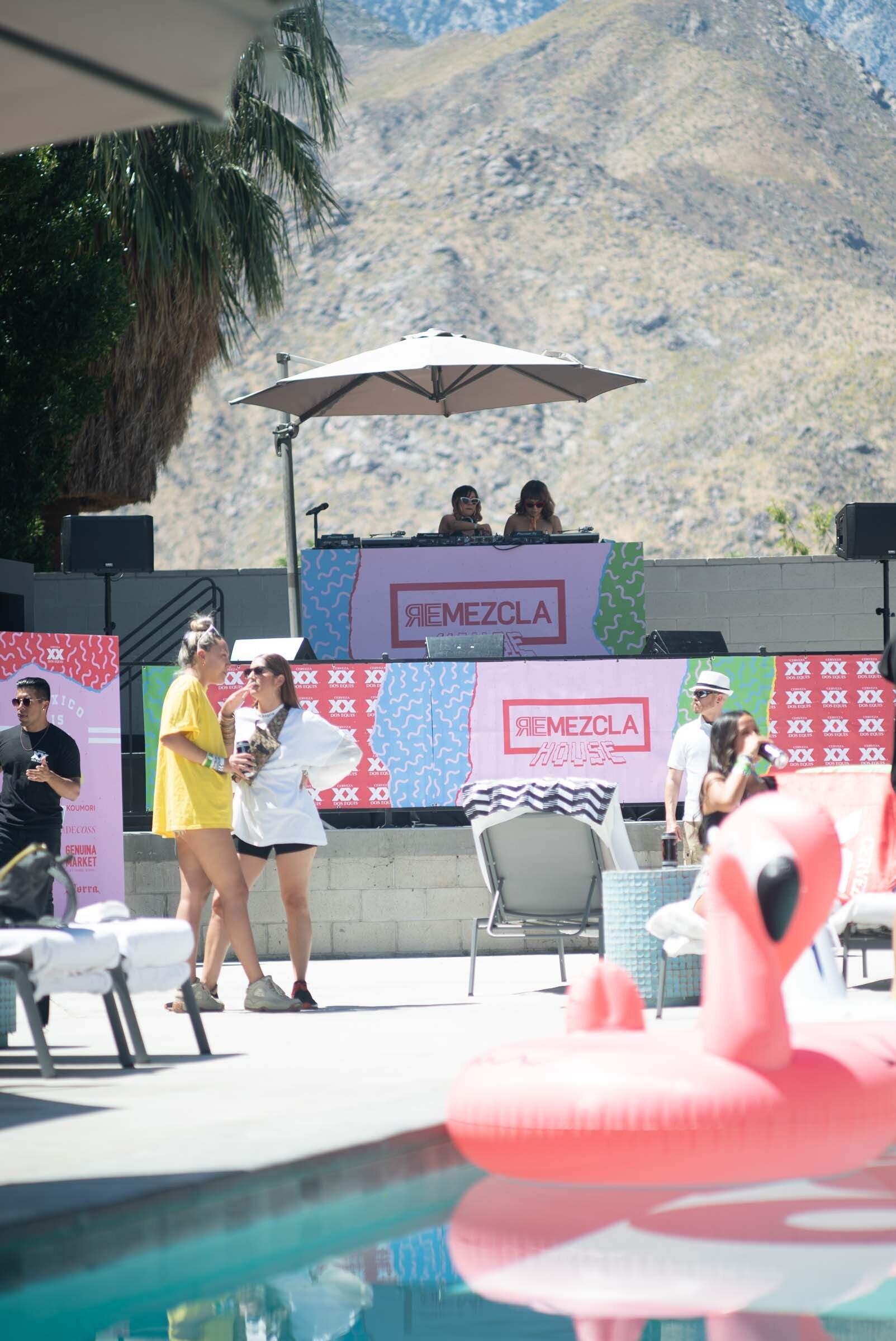 Personas en una fiesta junto a la piscina con un DJ en una plataforma elevada, palmeras y montañas de fondo, inflable de flamenco rosa en el agua.