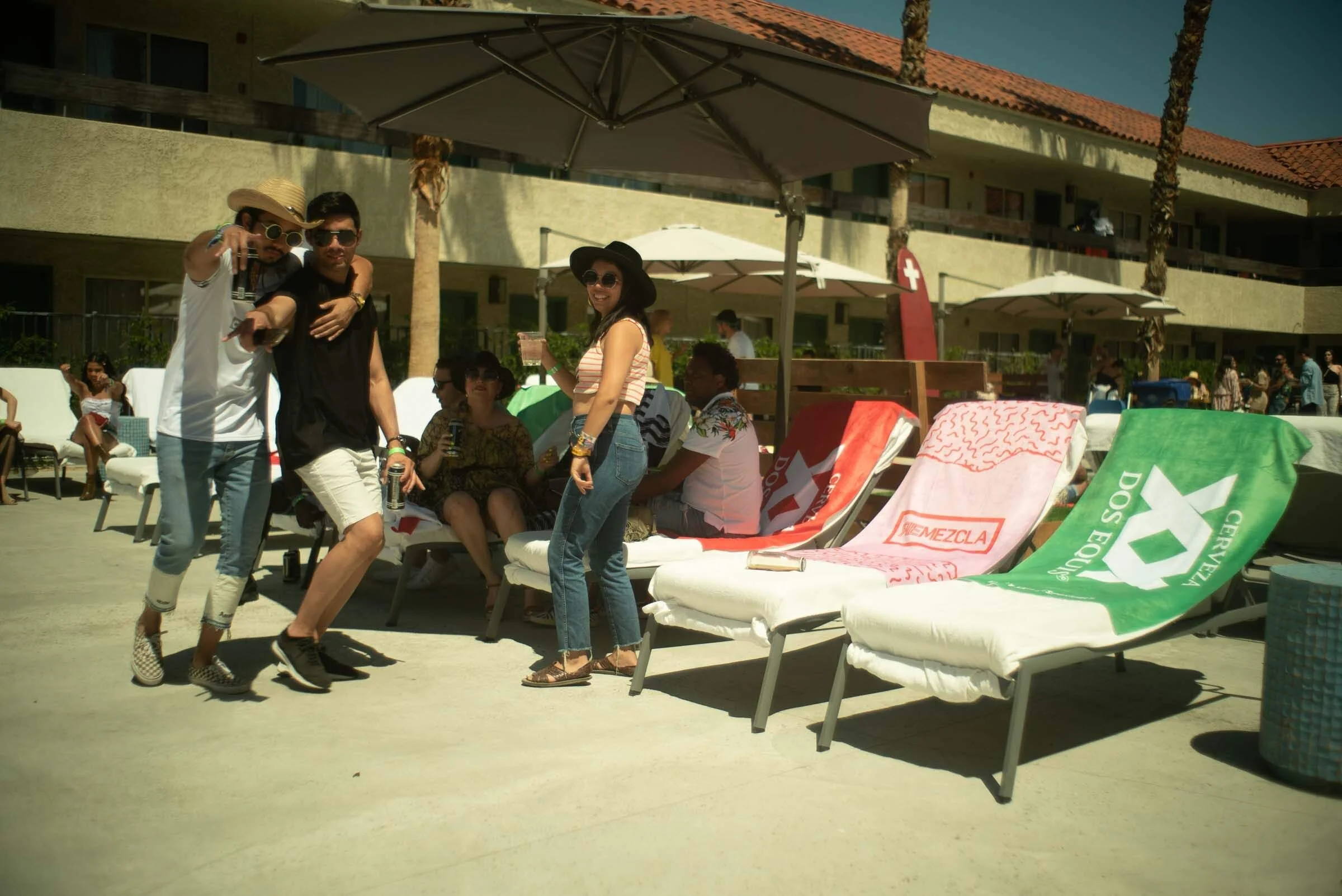 Grupo de personas disfrutando en una piscina al aire libre, algunos sentados en tumbonas con toallas y otros de pie con bebidas, en un día soleado en un entorno de resort o hotel con sombra de sombrillas y edificio al fondo.