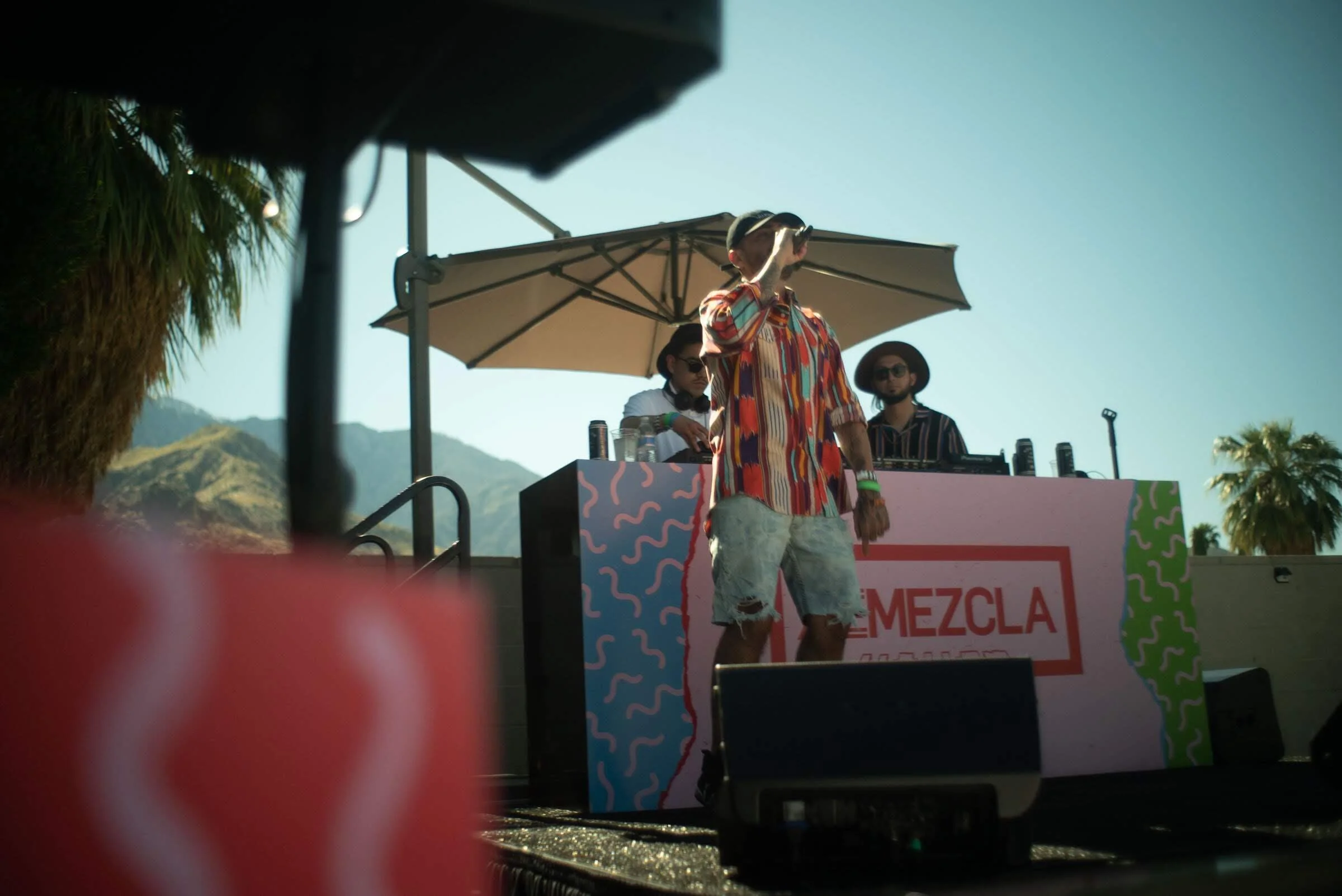 Tres personas en un escenario al aire libre, una de ellas con gafas y camiseta colorida, otra con gafas y sombrero, y una más con gafas y una camisa de rayas, con un cartel que dice 'Mezcla' y un paraguas, con montañas de fondo.
