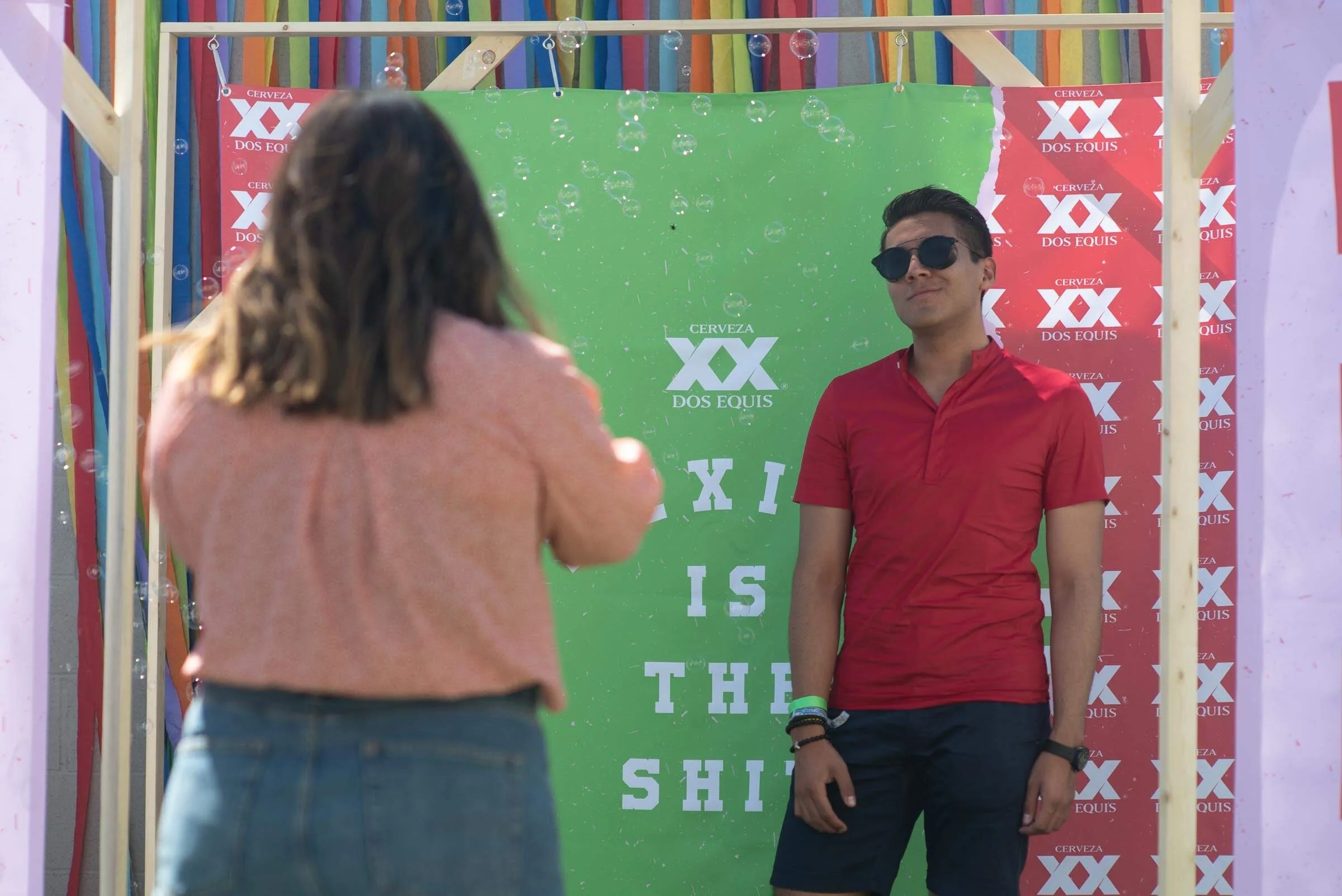 Una mujer toma una fotografía de un hombre que está frente a un fondo verde con el logo de Dos Equis y la frase "X is the Shit" en letras blancas. Ambos parecen estar en un evento o fotocabina decorada con banderas y globos.