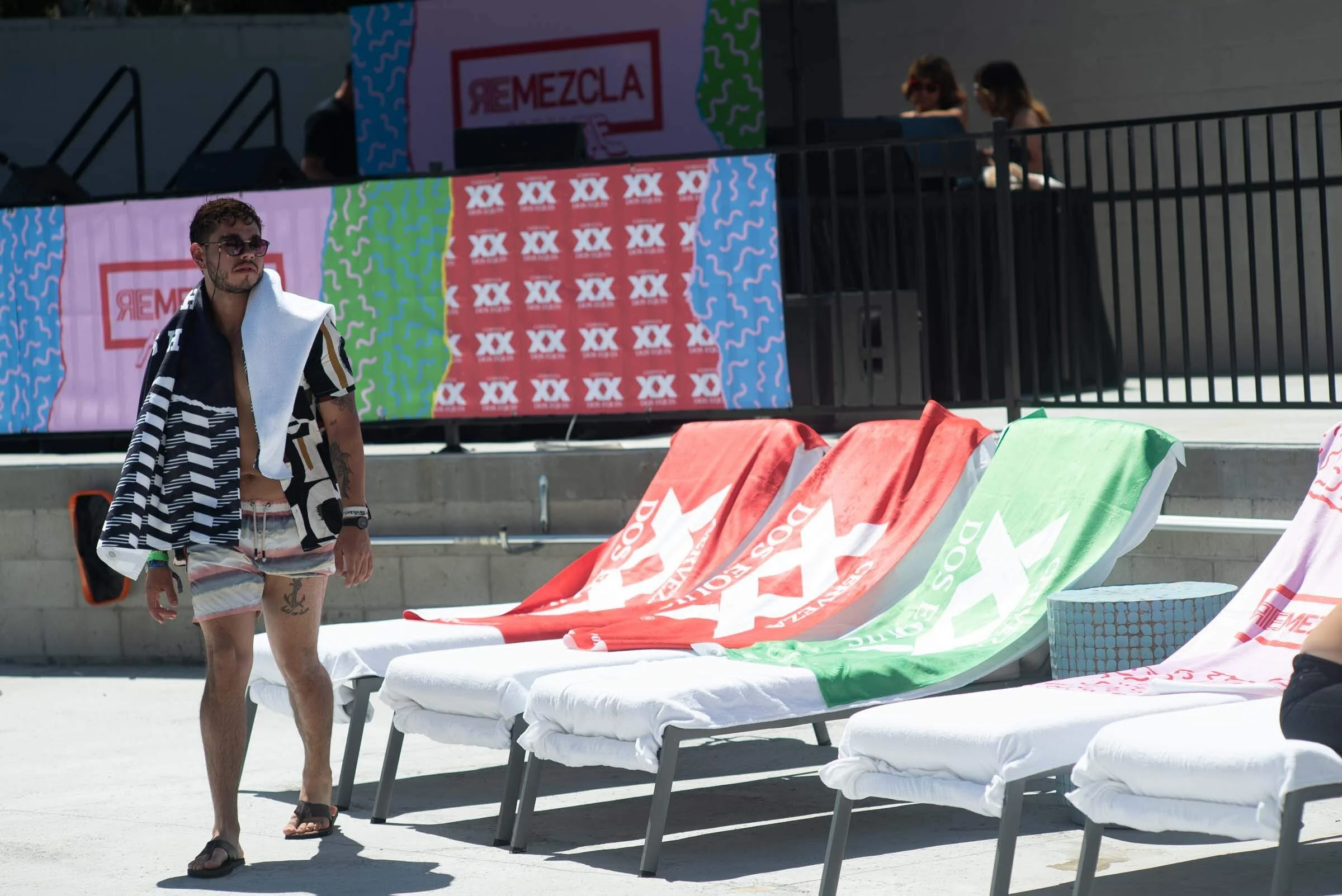 Un hombre con gafas, camisa abierta, short y sandalias, caminando junto a camastros con toallas y banderas de playa en un área de piscina, con pantallas digitales y dos mujeres en el fondo.
