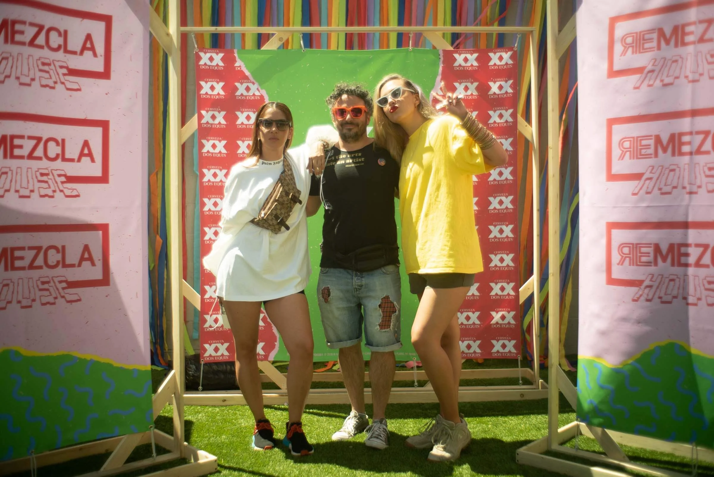 Tres personas posando juntas frente a un fondo con logotipos de cerveza y colores vibrantes en un evento al aire libre.