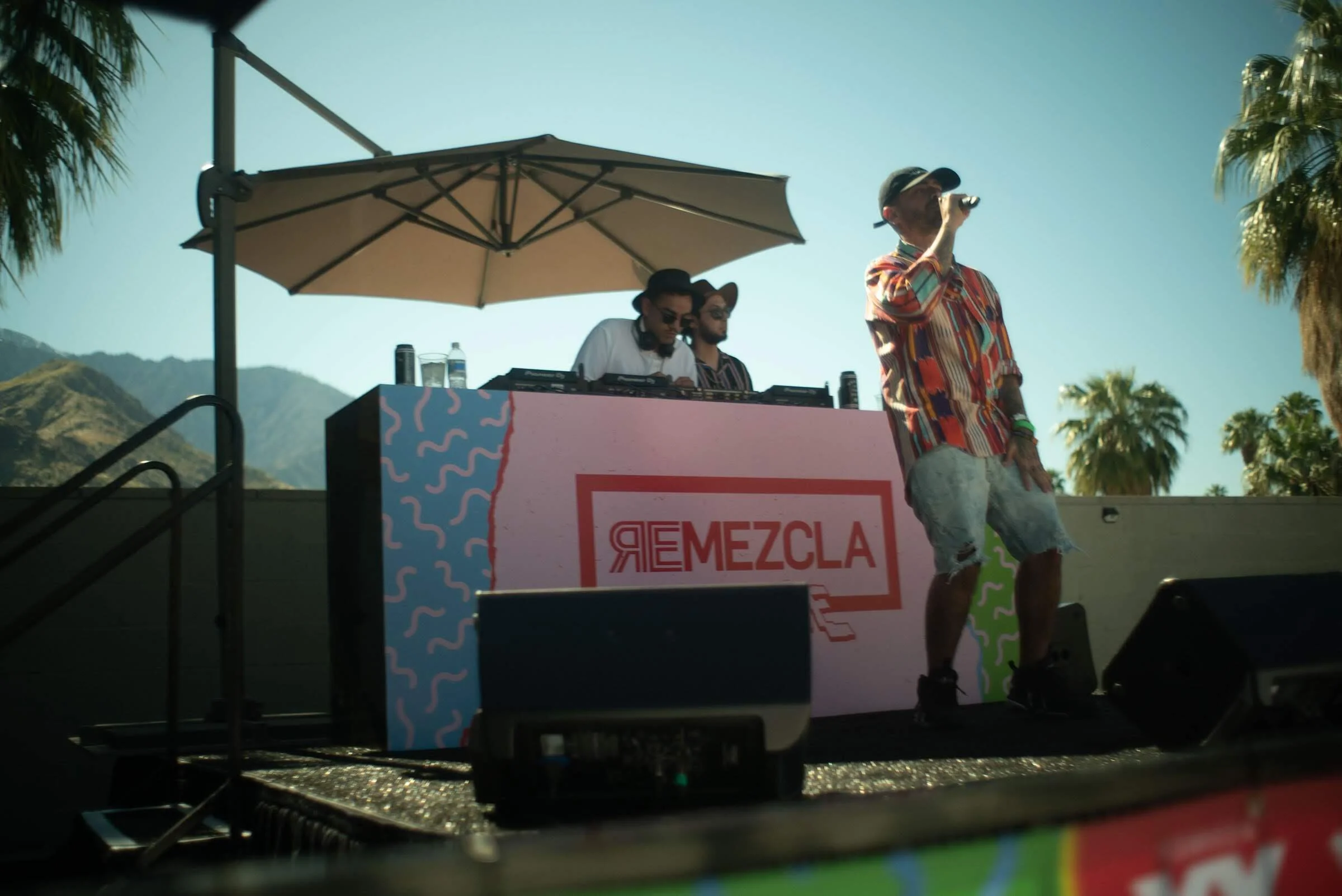Un DJ en un escenario al aire libre con una carpa y un paraguas, con montañas y palmeras en el fondo, y un cantante con micrófono vestido con una camisa a rayas y pantalones cortos delante del escenario.