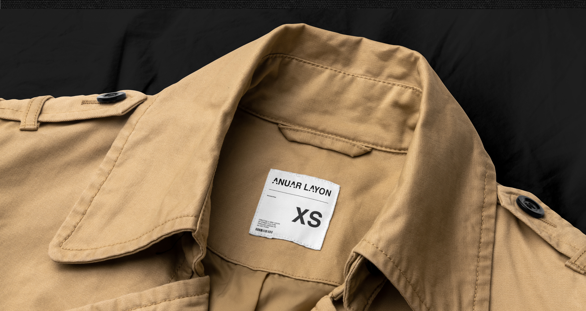 Vista cercana de la etiqueta de una chaqueta color café con la marca 'ANUAR LAYON' y tamaño XS, mostrando parte del cuello y botones en los hombros.