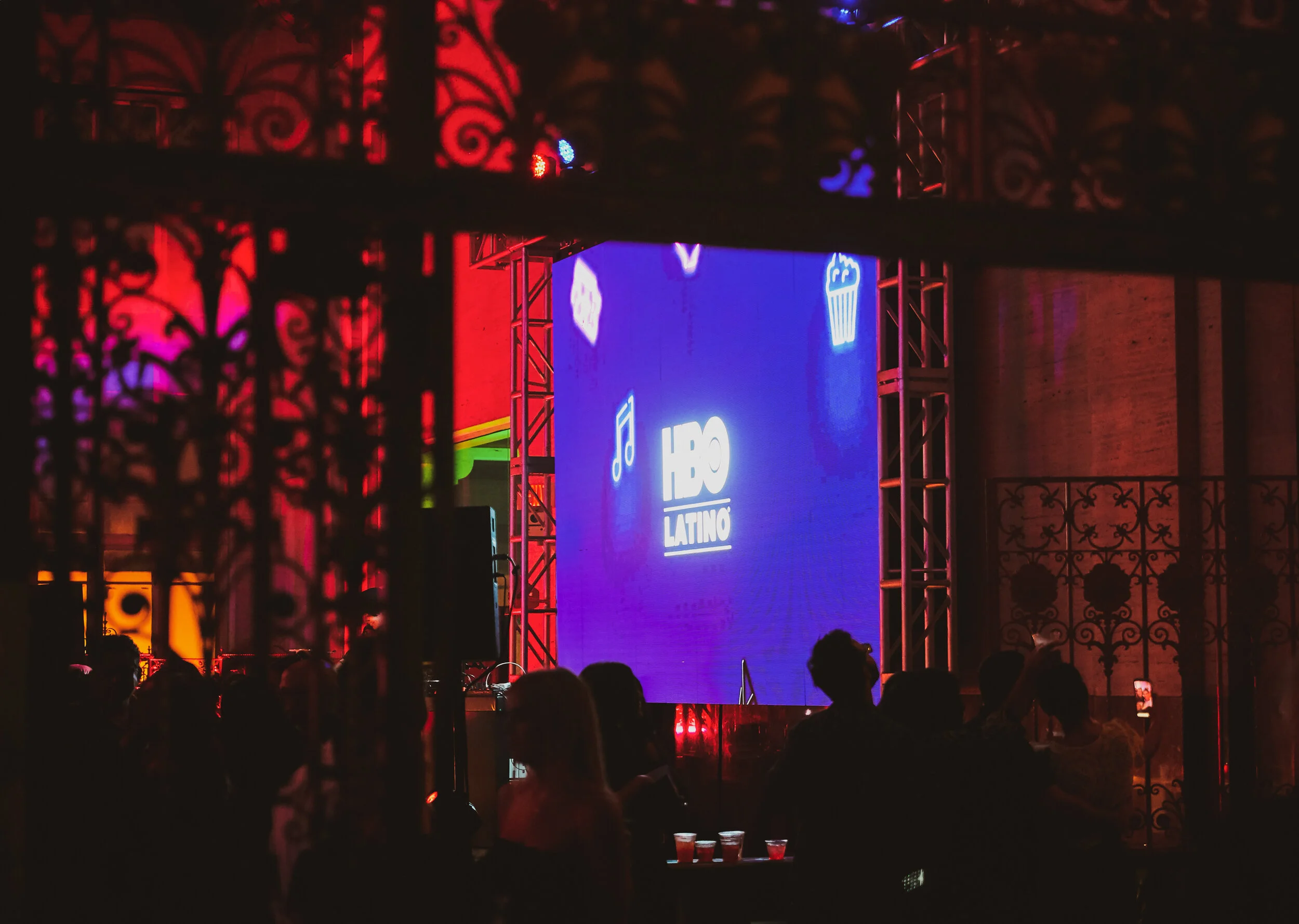 Escenario con pantalla grande que muestra el logo de HBO Latino y decoraciones con iconos de música, dulce y televisión, en un evento nocturno con varias personas en el público y mesas con bebidas.