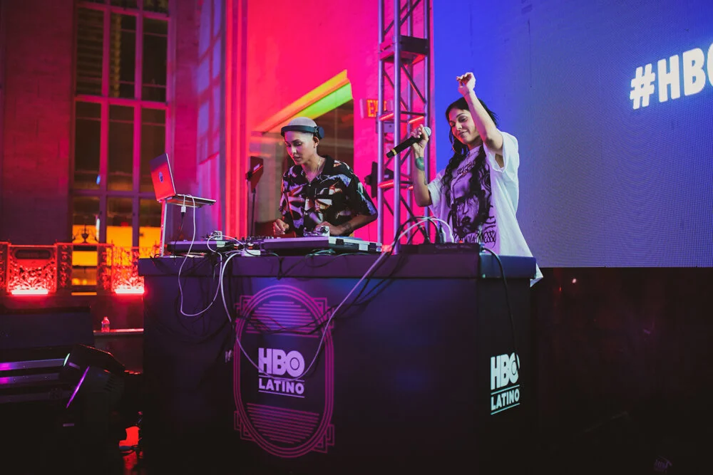 Dos personas en un escenario con decoraciones de luces, una tocando una consola de DJ y la otra con un micrófono, junto a un cartel de HBO Latino.