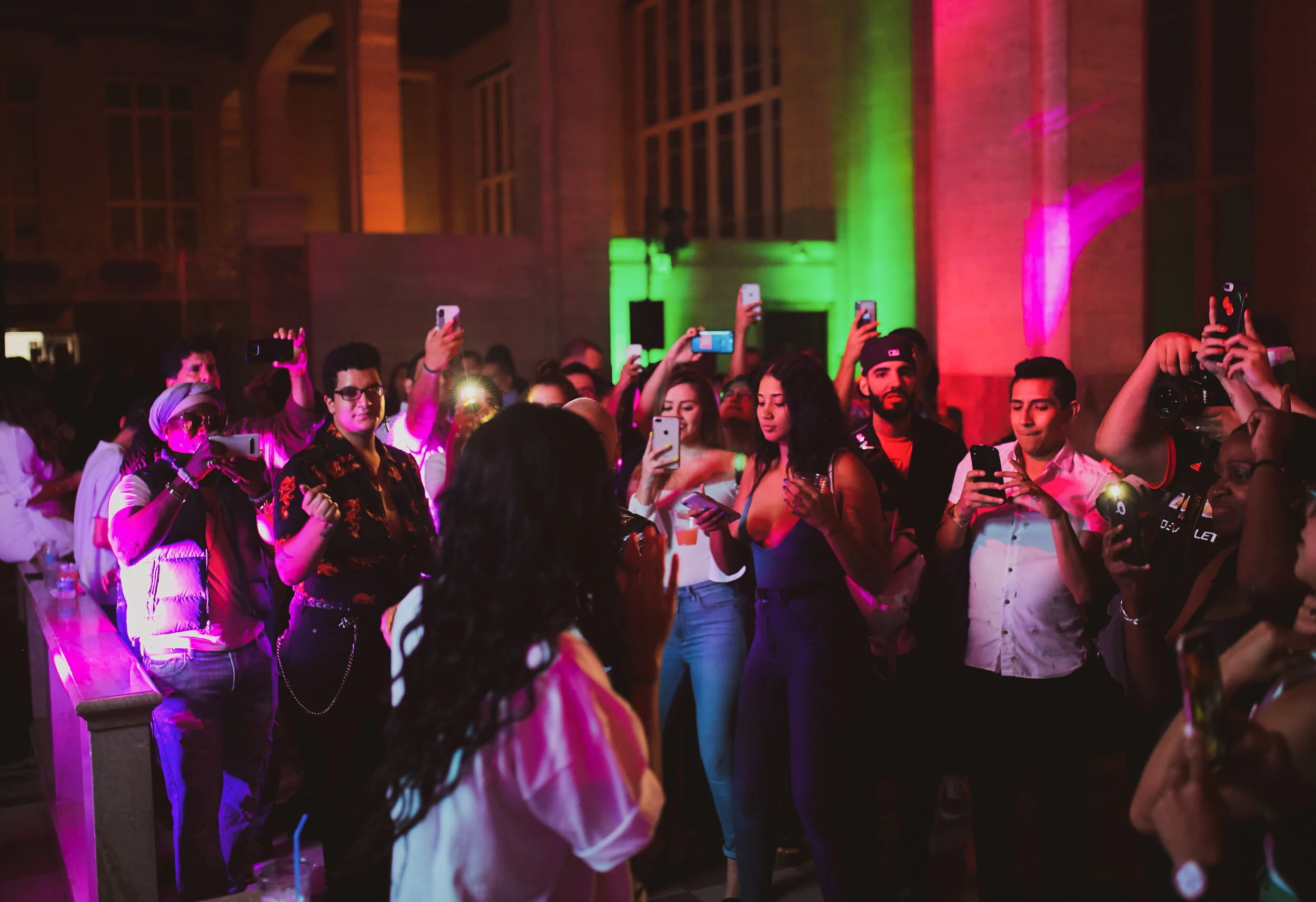 Personas en un evento nocturno con iluminación de colores, algunas usando lentes y tomándose fotos con teléfonos móviles.