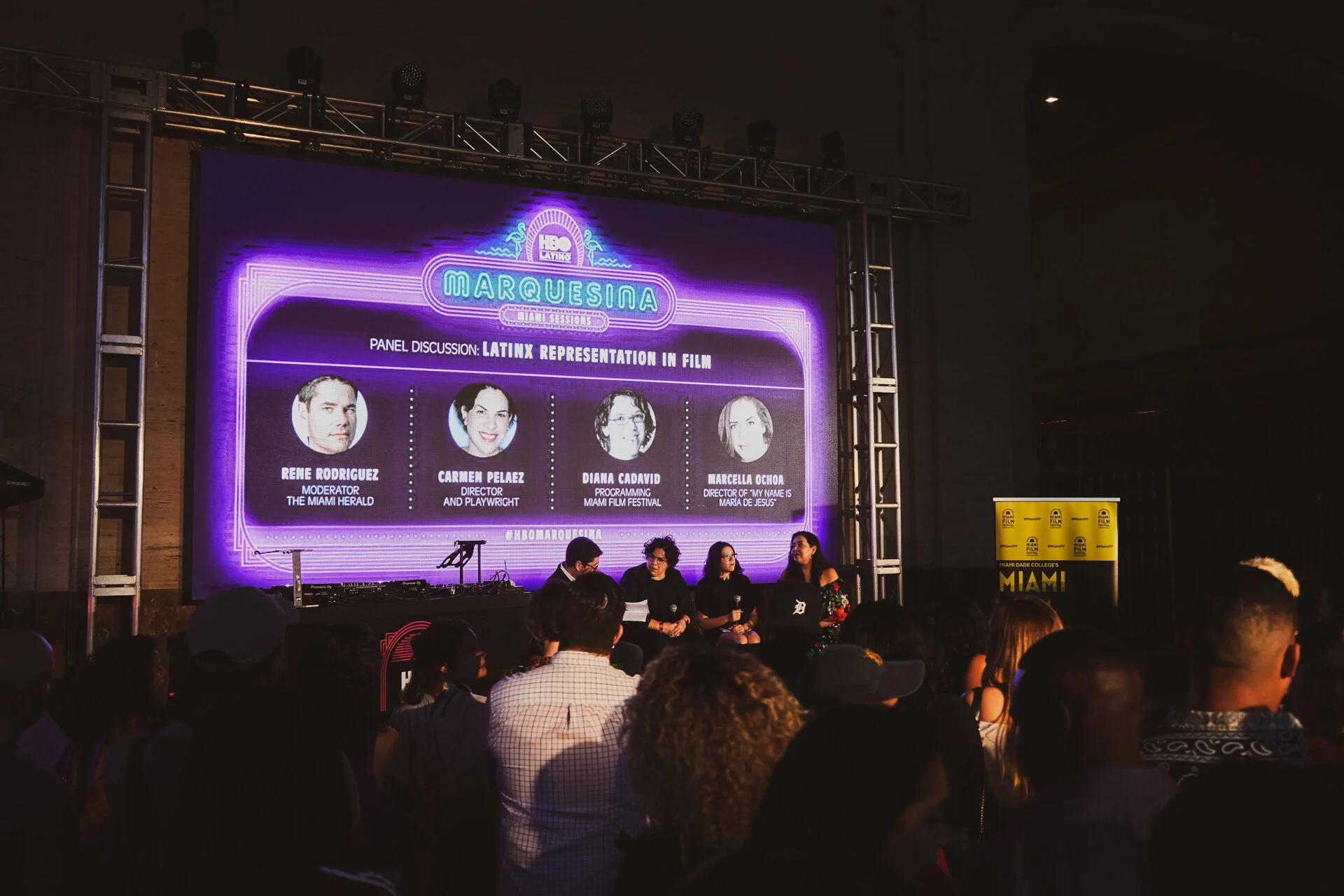 Panel de discusión en un evento de cine de temática latina, con cuatro panelistas en el escenario y una audiencia observando. La pantalla muestra información sobre la discusión y panelistas, en un fondo de colores morados y neón.