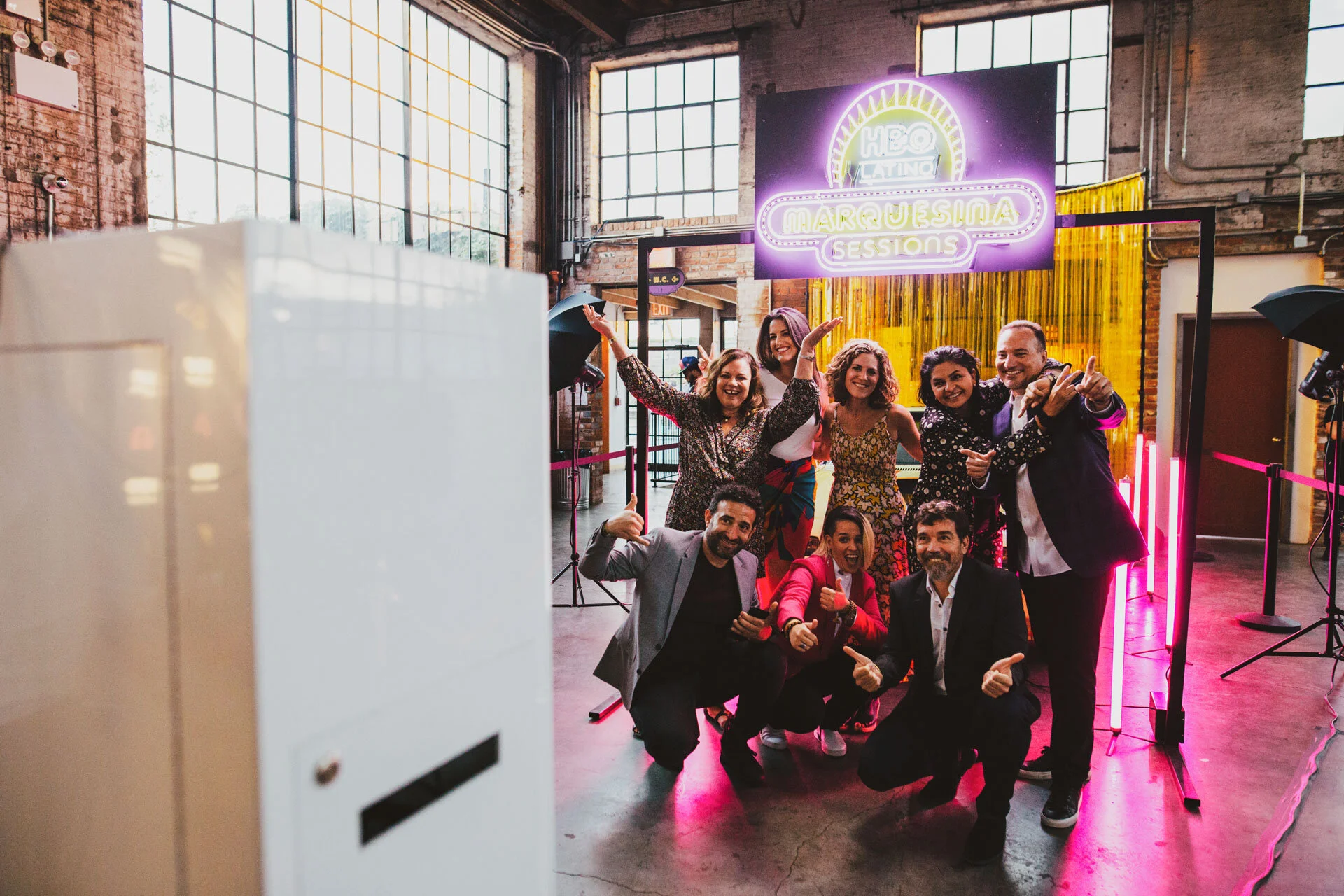 Grupo de personas sonrientes en un evento social, posando para la cámara con fondo de cartel de neón y decoración interior de ladrillo.