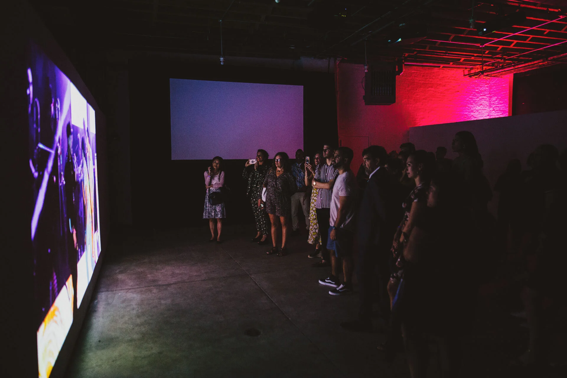 Grupo de personas observando una gran pantalla en una galería o museo con iluminación tenue y paredes con iluminación rosa.