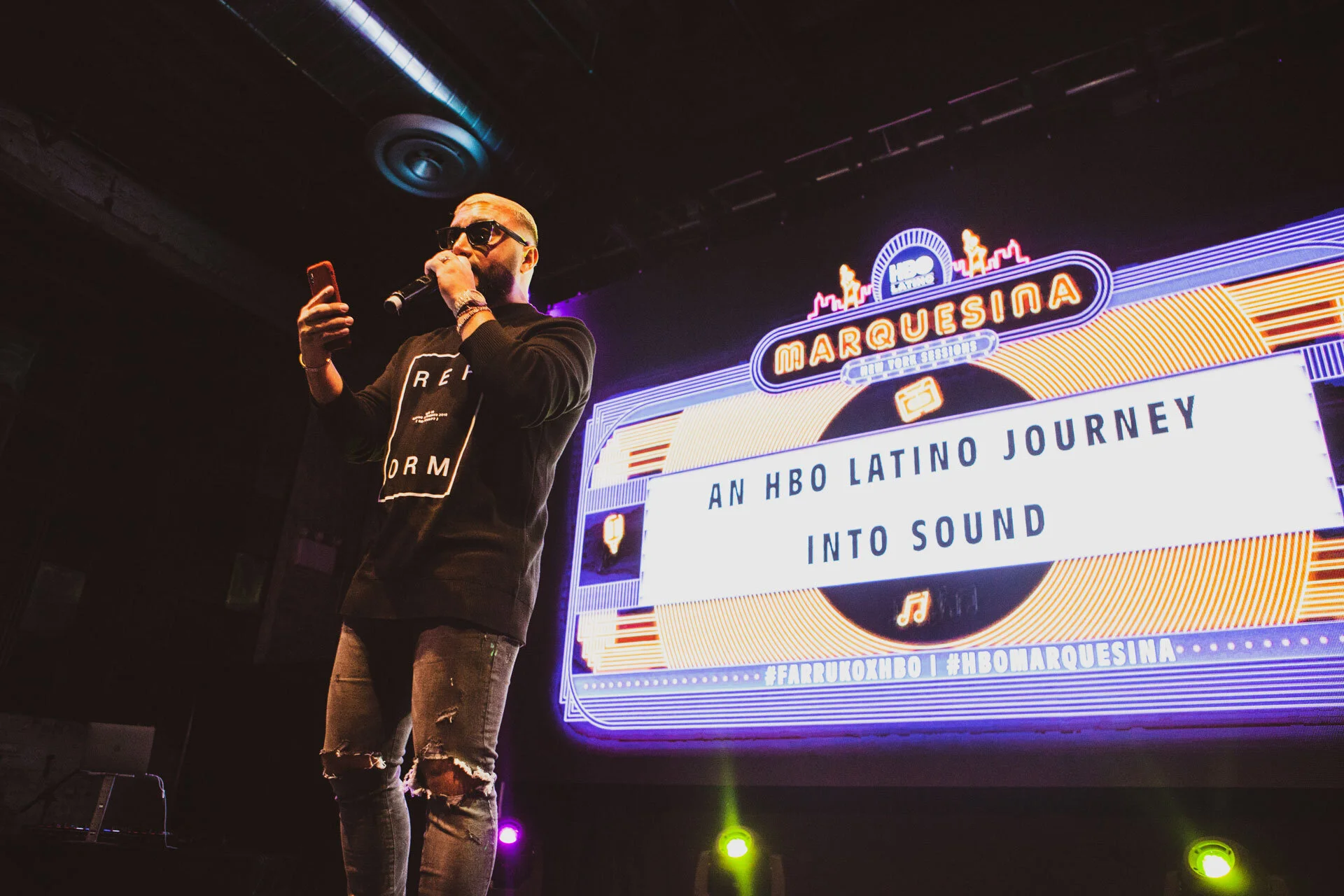 Un hombre de pie en un escenario, sosteniendo un micrófono y un teléfono celular, con una pantalla grande detrás de él que dice 'HBO MarQueSina' y el texto 'Un viaje latino de HBO hacia el sonido'.