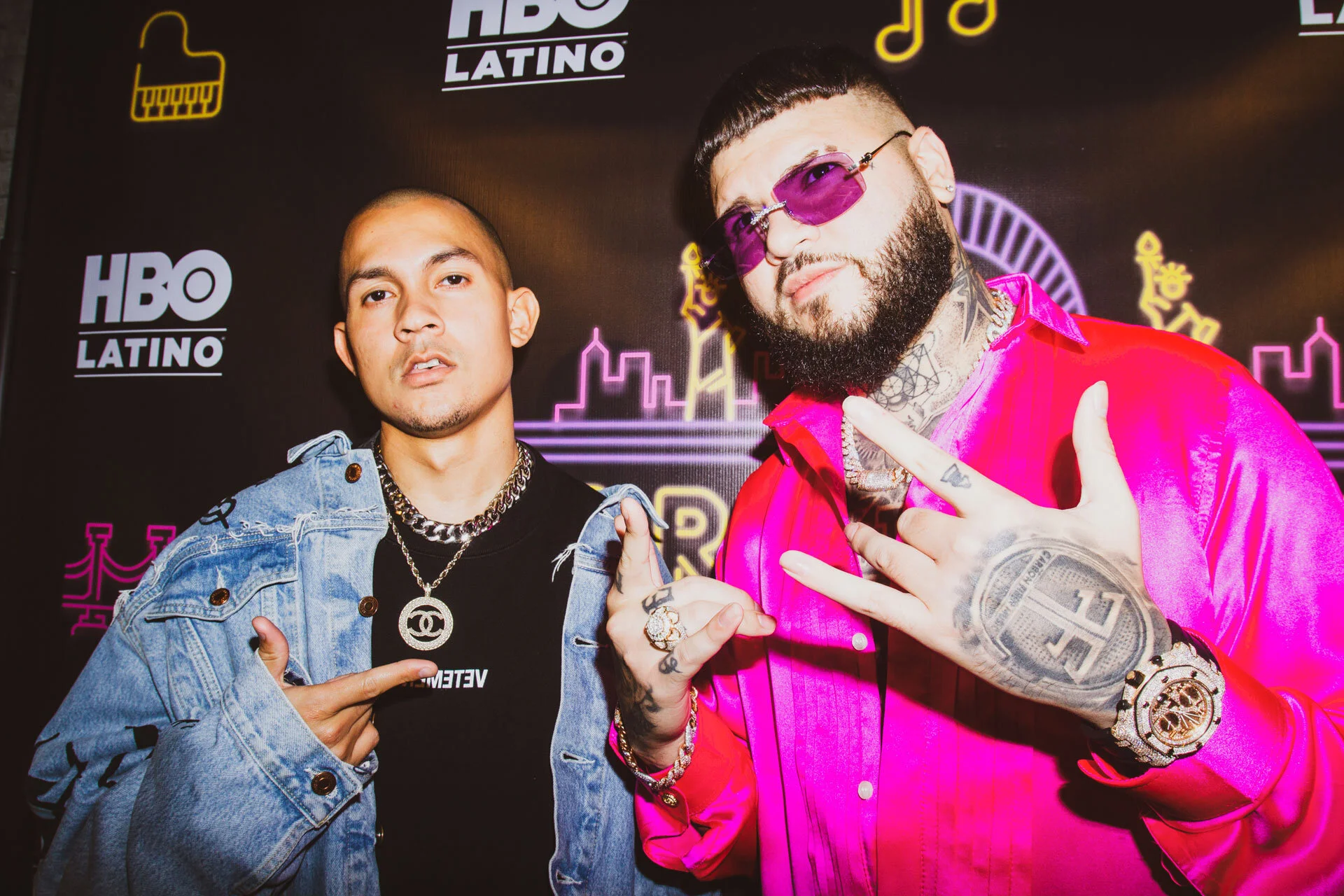 Dos hombres en un evento de HBO Latino. Uno lleva una chaqueta de mezclilla y cadenas grandes, señalando con su mano. El otro lleva gafas rosadas y una camisa rosa brillante, mostrando una señal de mano. Fondo con logotipos de HBO Latino y neon de un