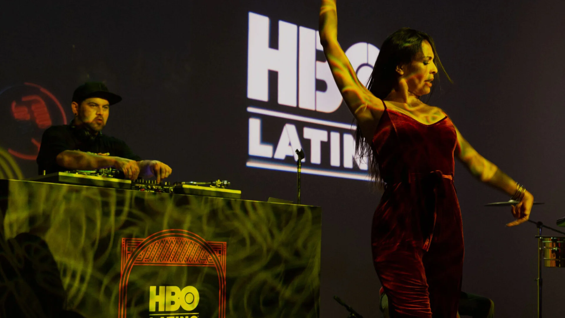 Un DJ y una bailarina en un escenario con logotipo de HBO, la bailarina viste un vestido rojo y está en una pose con un brazo levantado.