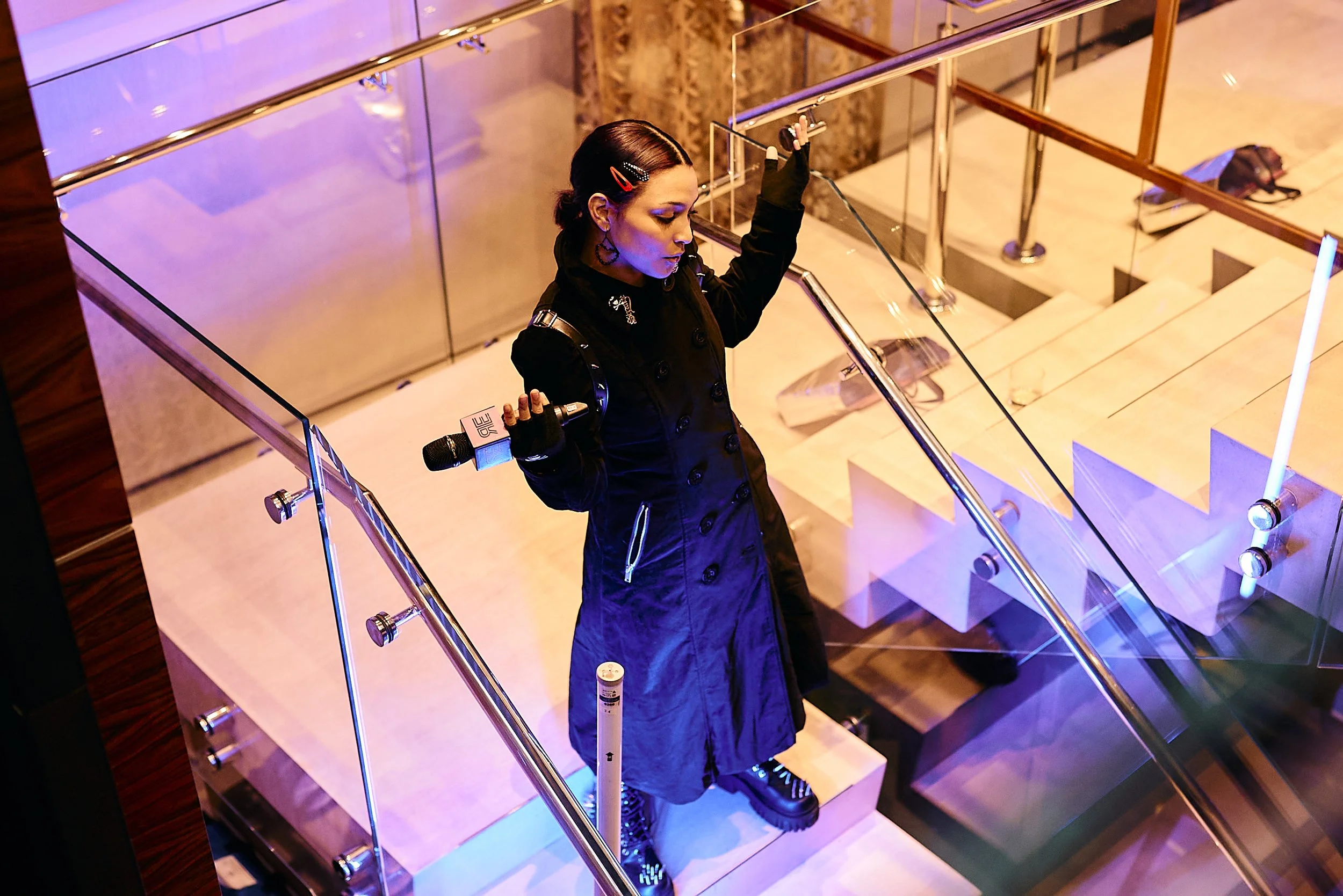Una mujer joven con estilo alternativo y maquillaje oscuro, vestida con un abrigo largo negro, está en las escaleras del interior de un edificio moderno, sosteniendo un micrófono en una mano y la barandilla con la otra, con iluminación de luces de co