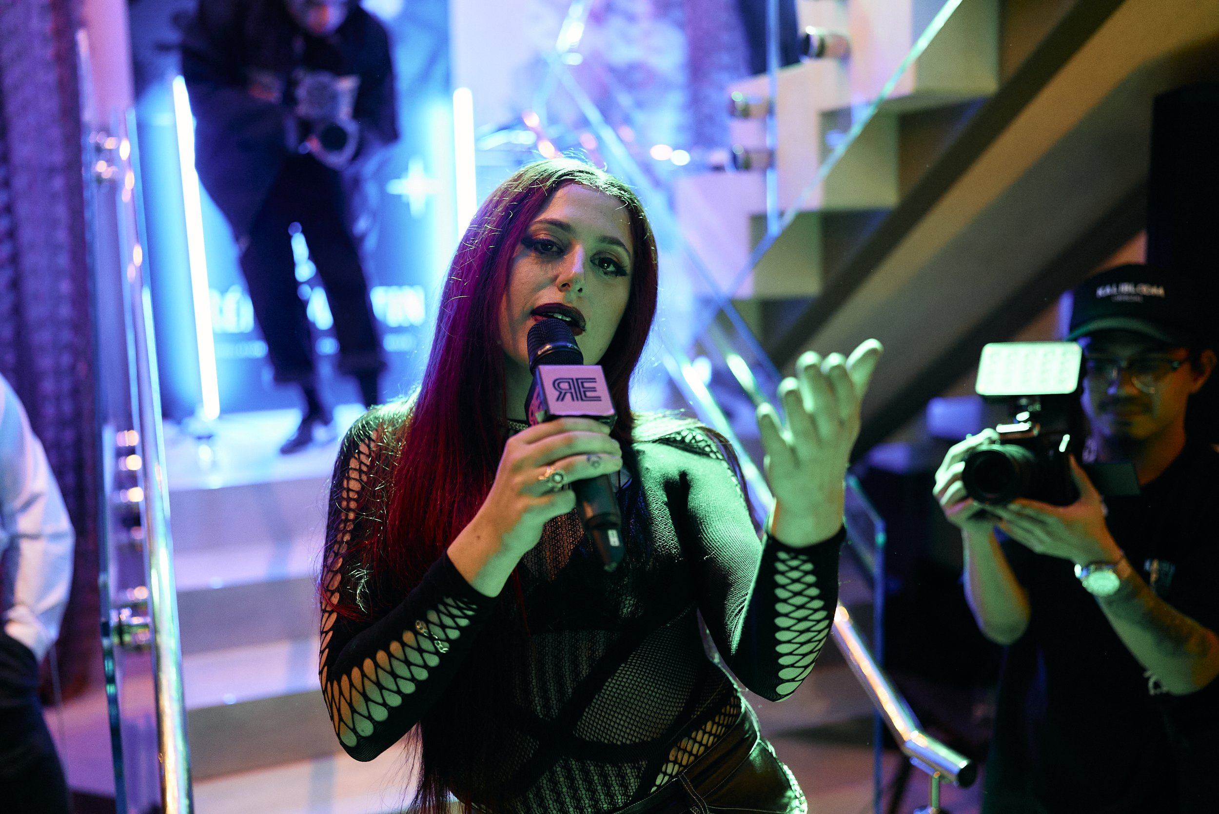 Una mujer con cabello rojo y ropa negra con mallas de red sostiene un micrófono en un evento con iluminación de colores. Hay un hombre con gorra y un cámara grabando.