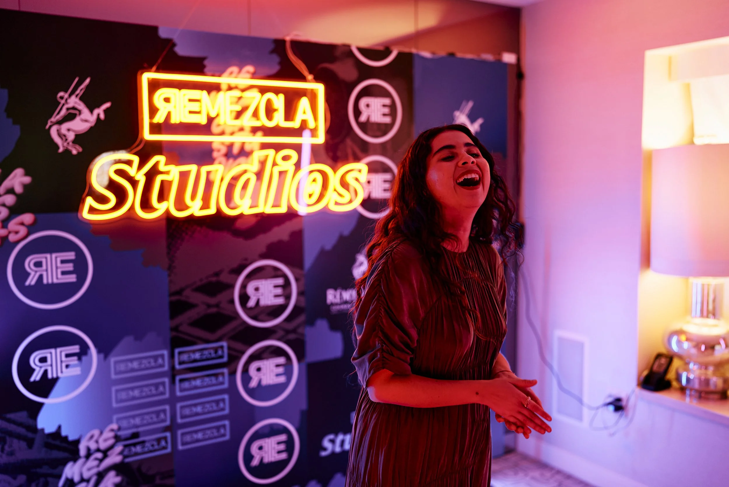 Una mujer sonriendo y riendo en un estudio con un letrero de neón que dice 'REMEZCLA Studios' en el fondo, con paredes decoradas y una lámpara al lado.
