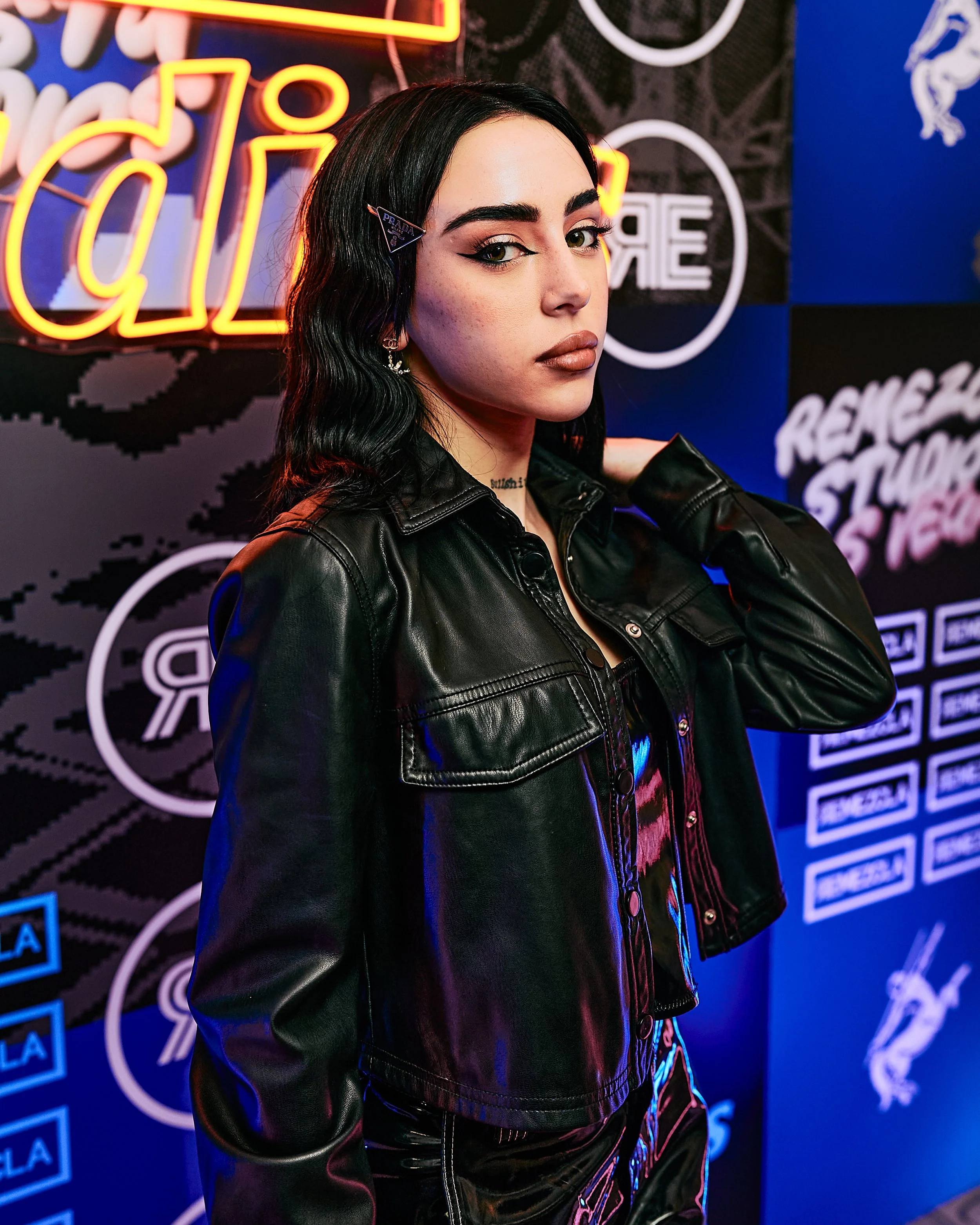 Joven mujer con cabello negro y piel clara en un ambiente con luces neón, lleva chaqueta de cuero negra y está posando en un fondo con logotipos y neón