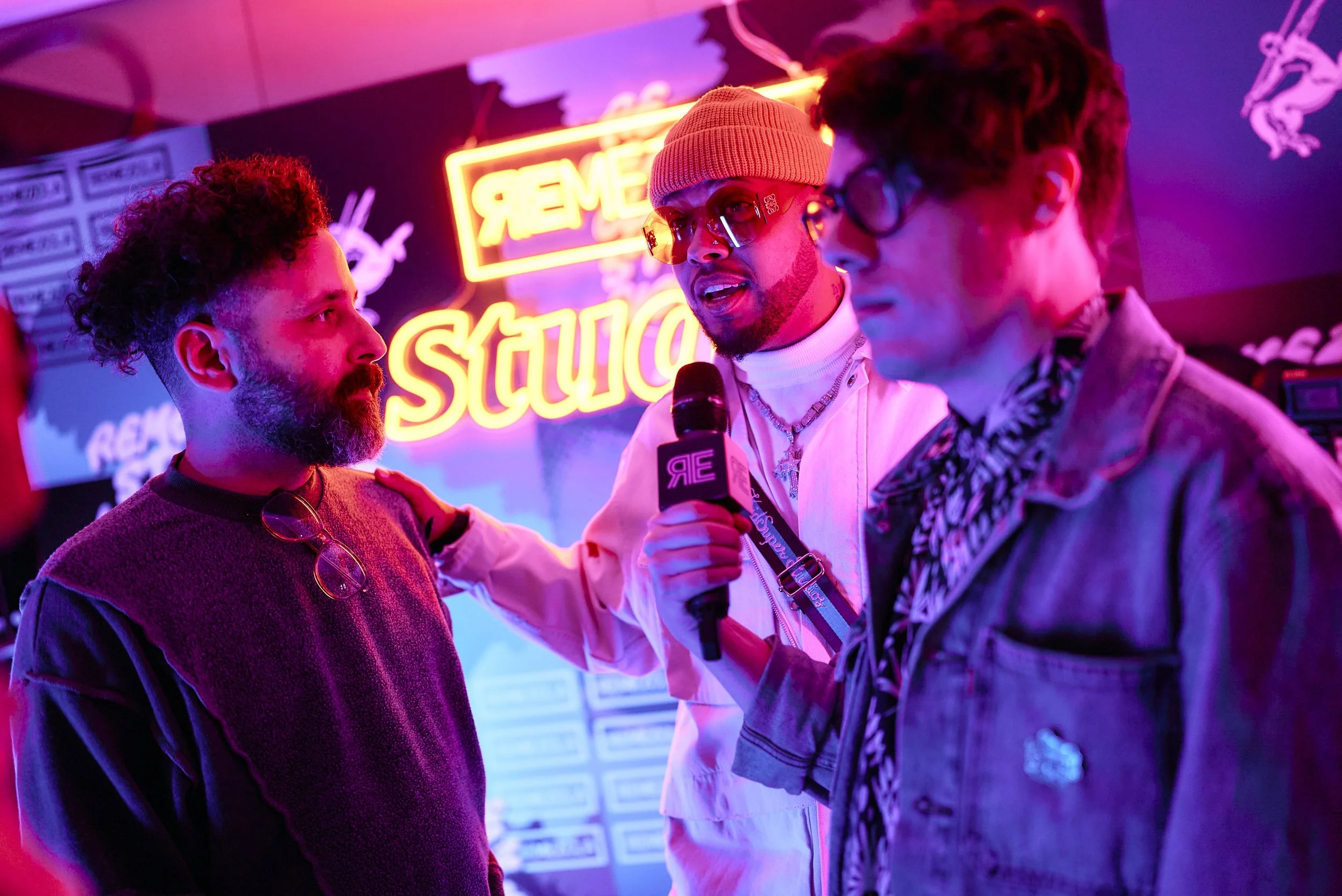 Tres personas en un fondo con neón que dice 'STUDENT' en un ambiente con luces coloridas, una de ellas sostiene un micrófono.