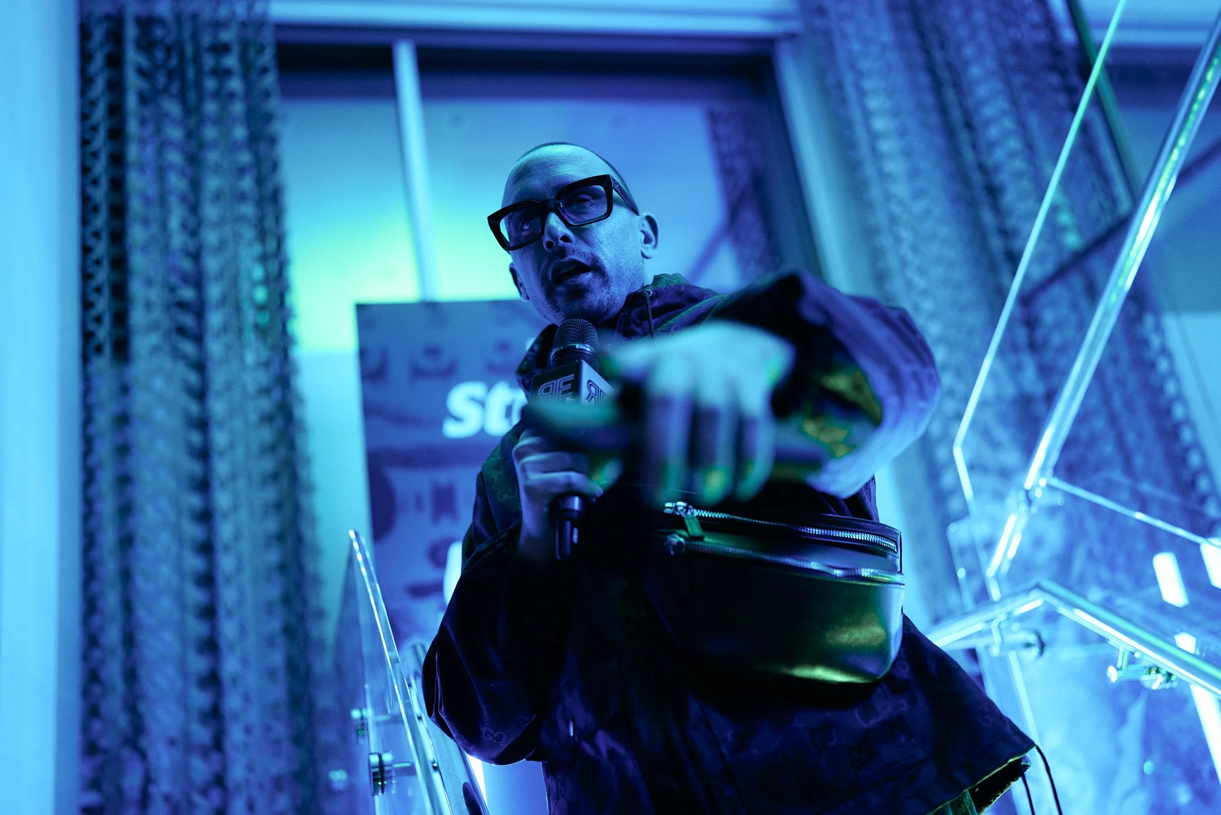 Hombre con gafas y chaqueta sosteniendo un micrófono, señalando hacia la cámara, en un escenario con iluminación azul y fondo con cortinas y luces LED.