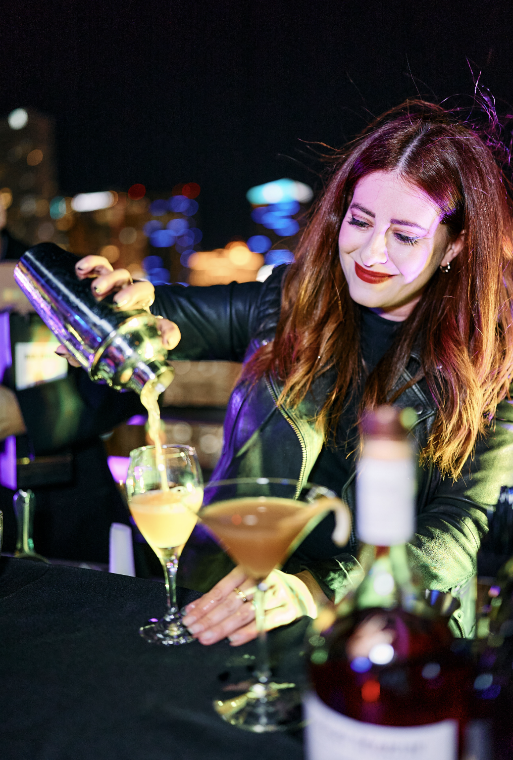 Mujer con cabello rizado y maquillaje rojo sirviendo una bebida en un cóctel en un bar con luces de ciudad al fondo.
