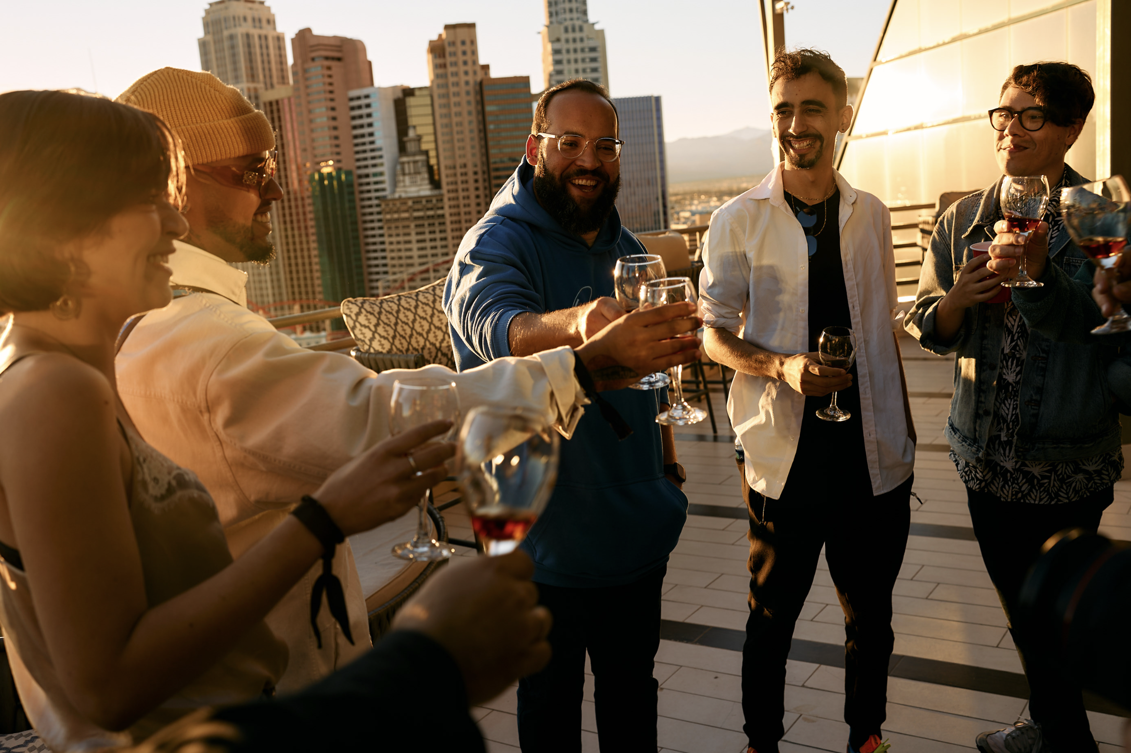 Grupo de personas brindando con copas de vino en una terraza con vista a la ciudad al atardecer.