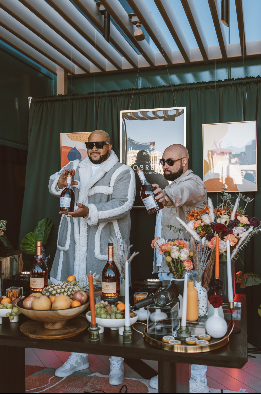 Dos hombres en una celebración con mesas llenas de fruta, vino y flores, sosteniendo botellas de alcohol, en un espacio decorado con cuadros y plantas.