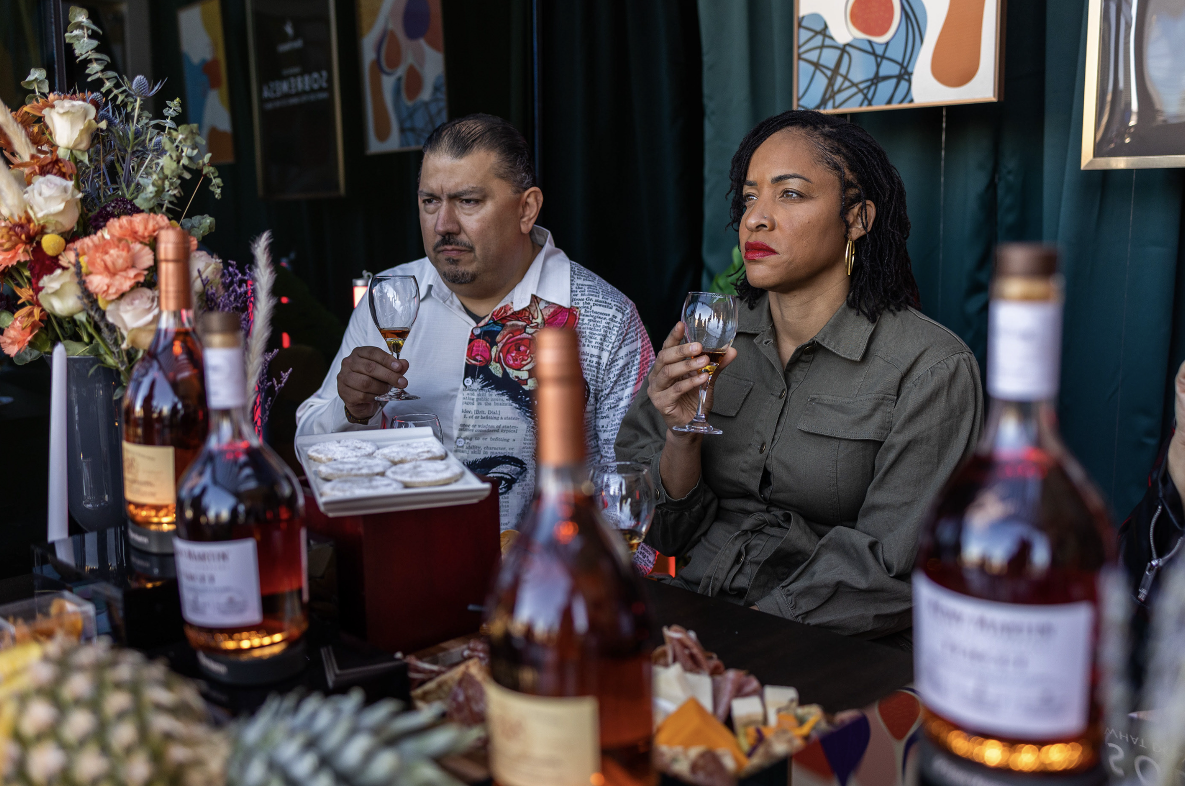 Dos personas sentadas en una mesa con bebidas y bocadillos, observando con expresión seria, en un ambiente con decoraciones florales y cuadros en la pared.