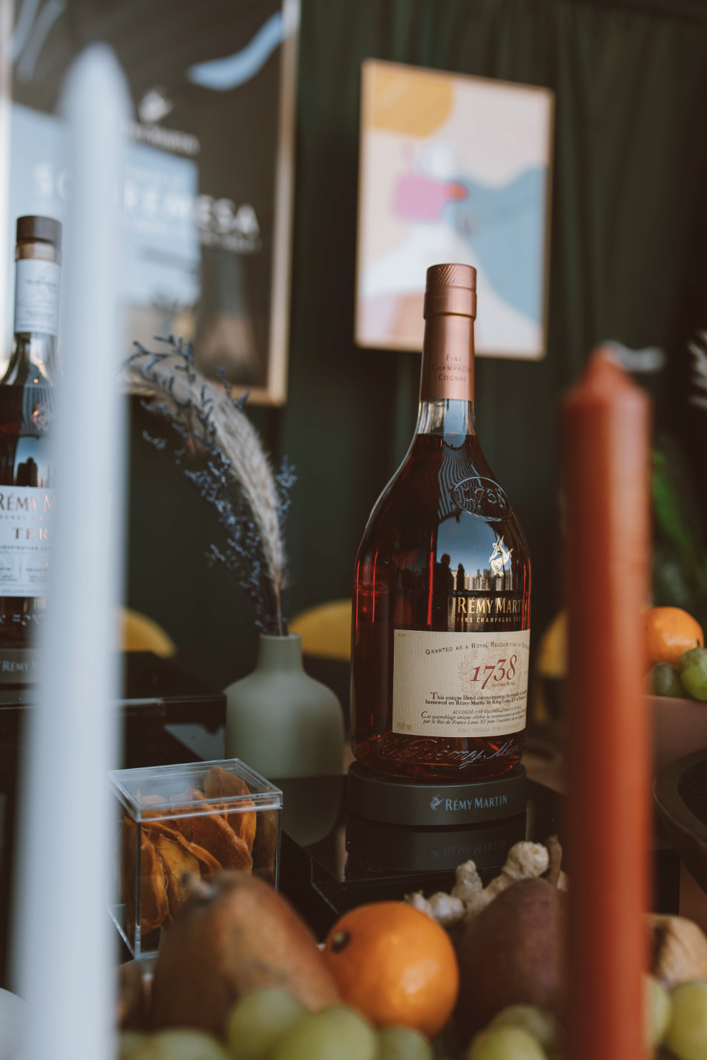 Botella de coñac Rémy Martin en un entorno decorativo con frutas, una vela y una flor seca en un jarrón, en un espacio que parece ser una exhibición o tienda de bebidas.