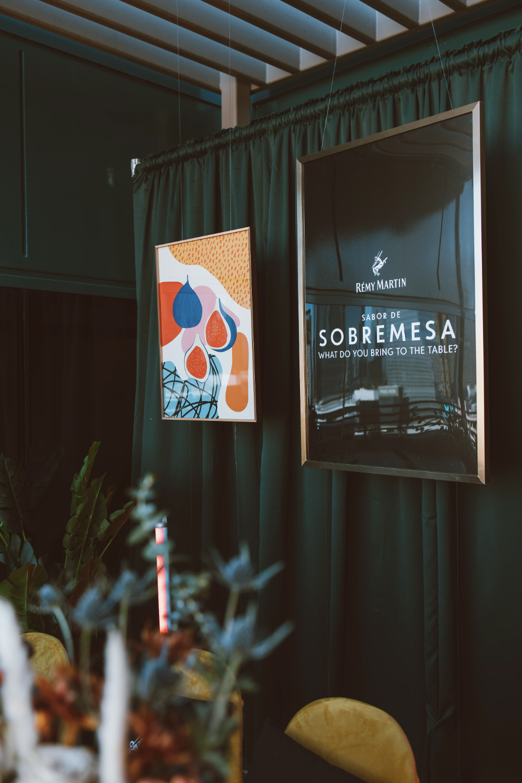 Carteles en una pared con cortinas, uno con arte abstracto de frutas y otro con texto promocional de Remy Martin sobre el sabor de sobremesa.