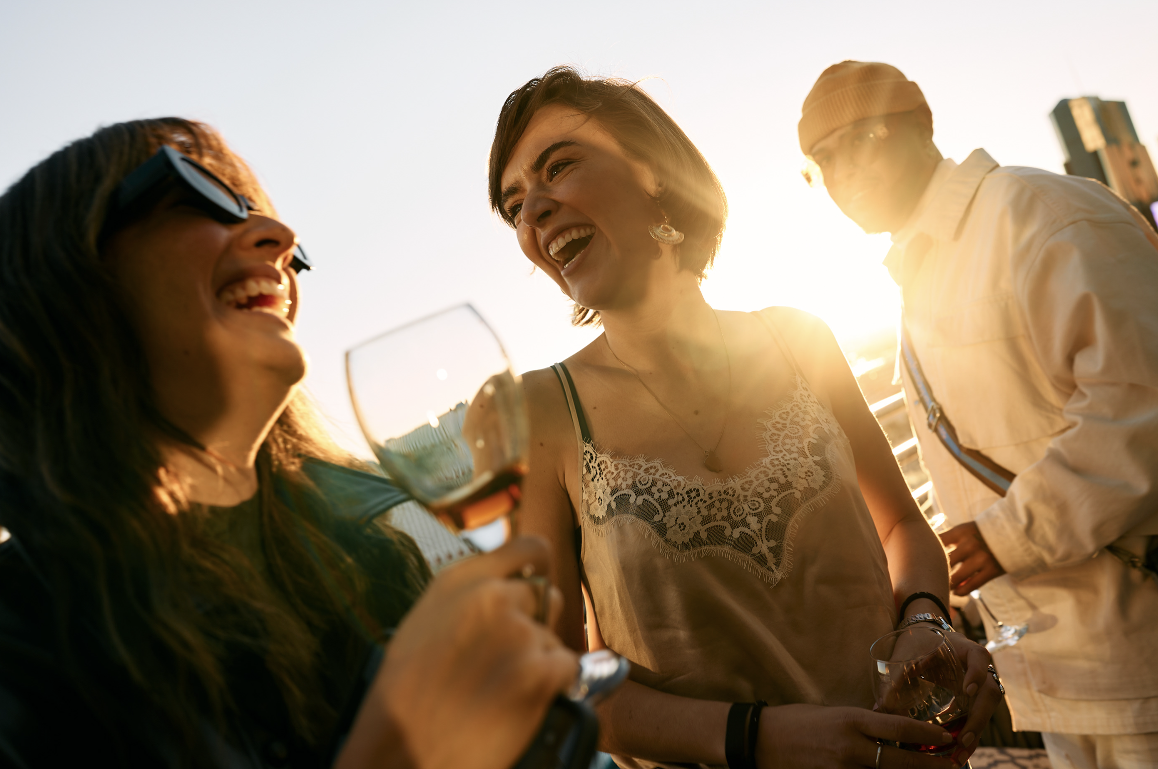 Tres personas jóvenes disfrutando en una terraza al atardecer, riendo y conversando, una mujer con gafas de sol y una copa de vino en mano, otra mujer con camiseta de encaje y un hombre con gorra y gafas, en un ambiente relajado y alegre.
