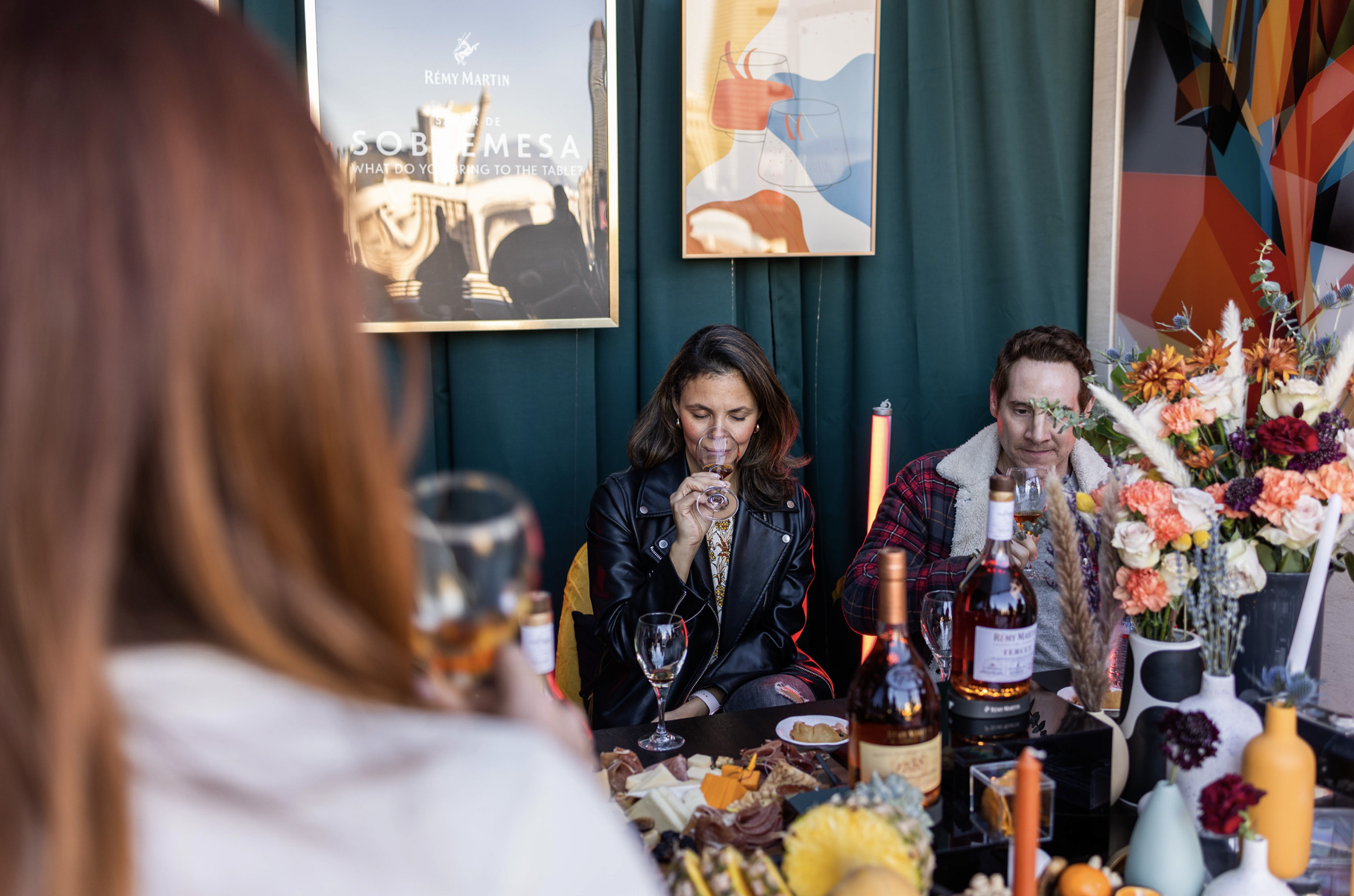 Personas apreciando una degustación con vino y queso en un ambiente decorado con flores y arte en las paredes.
