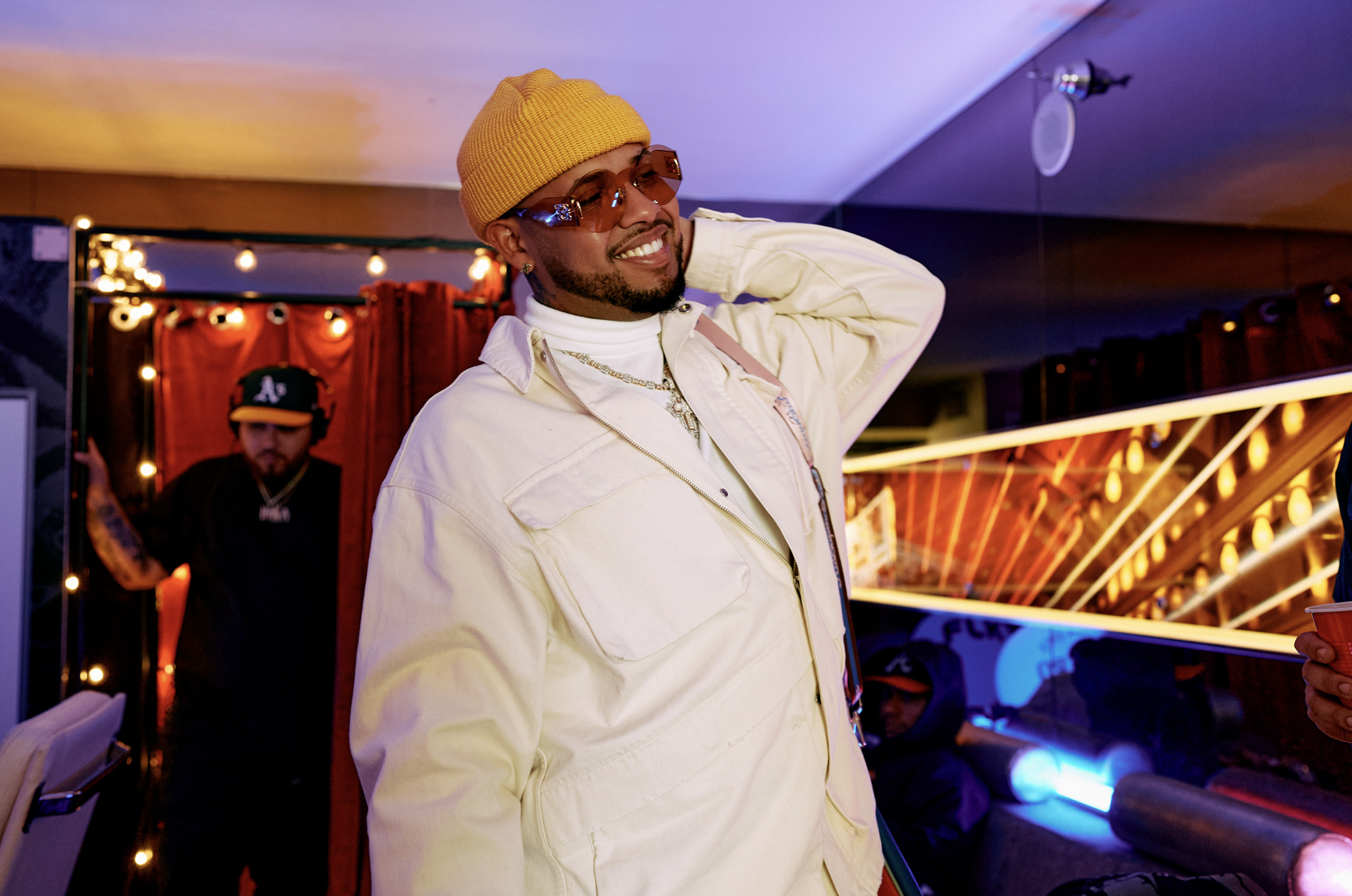 Hombre sonriendo con gafas de sol, gorra amarilla y ropa blanca en una fiesta con luces y otras personas en el fondo.