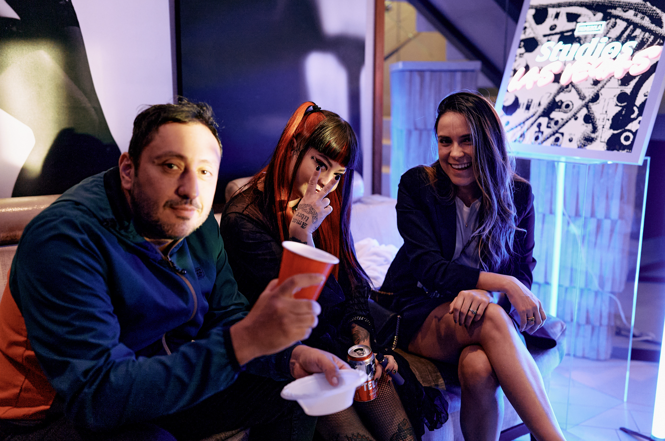 Tres personas sentadas en un sillón en un ambiente con luces de neón y un cartel que dice 'estudios en Texas', disfrutando de bebidas y sonriendo.