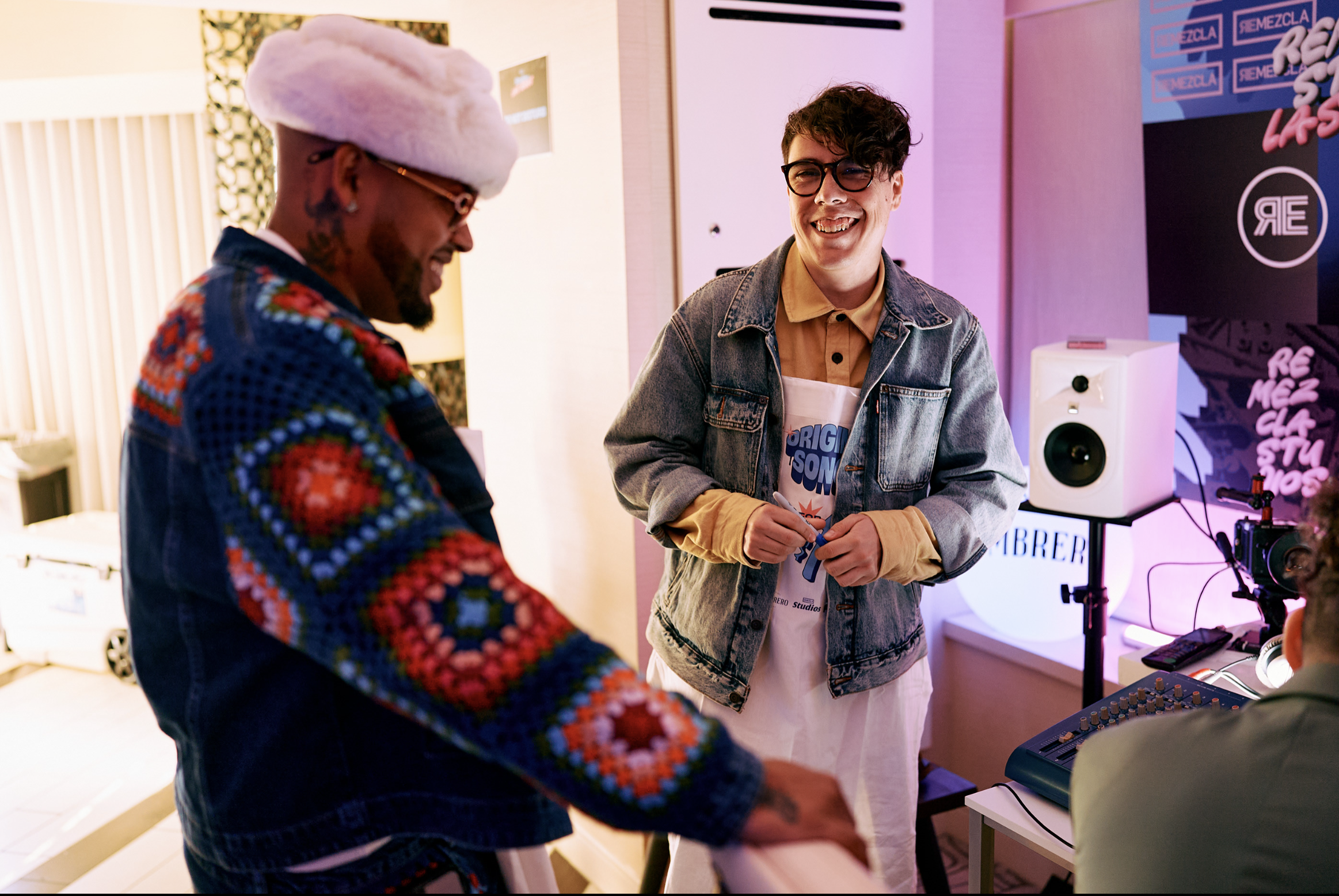 Dos hombres en un estudio de grabación, uno sonriendo y el otro con la cabeza abajo, rodeados de equipo de sonido y con un cartel que dice 'REMEZCLA'.