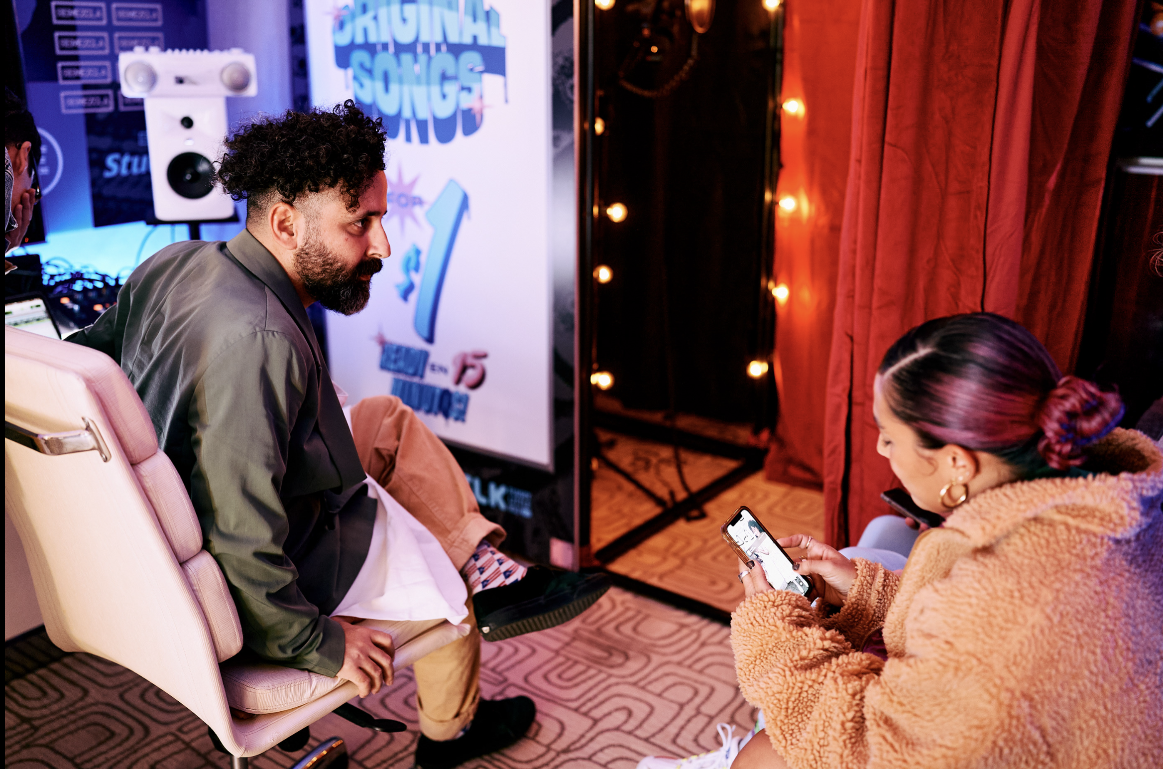 Una mujer con abrigo de peluche anaranjado usando un teléfono, sentada frente a un hombre con cabello rizado y barba, en un ambiente con luces y cortinas rojas.