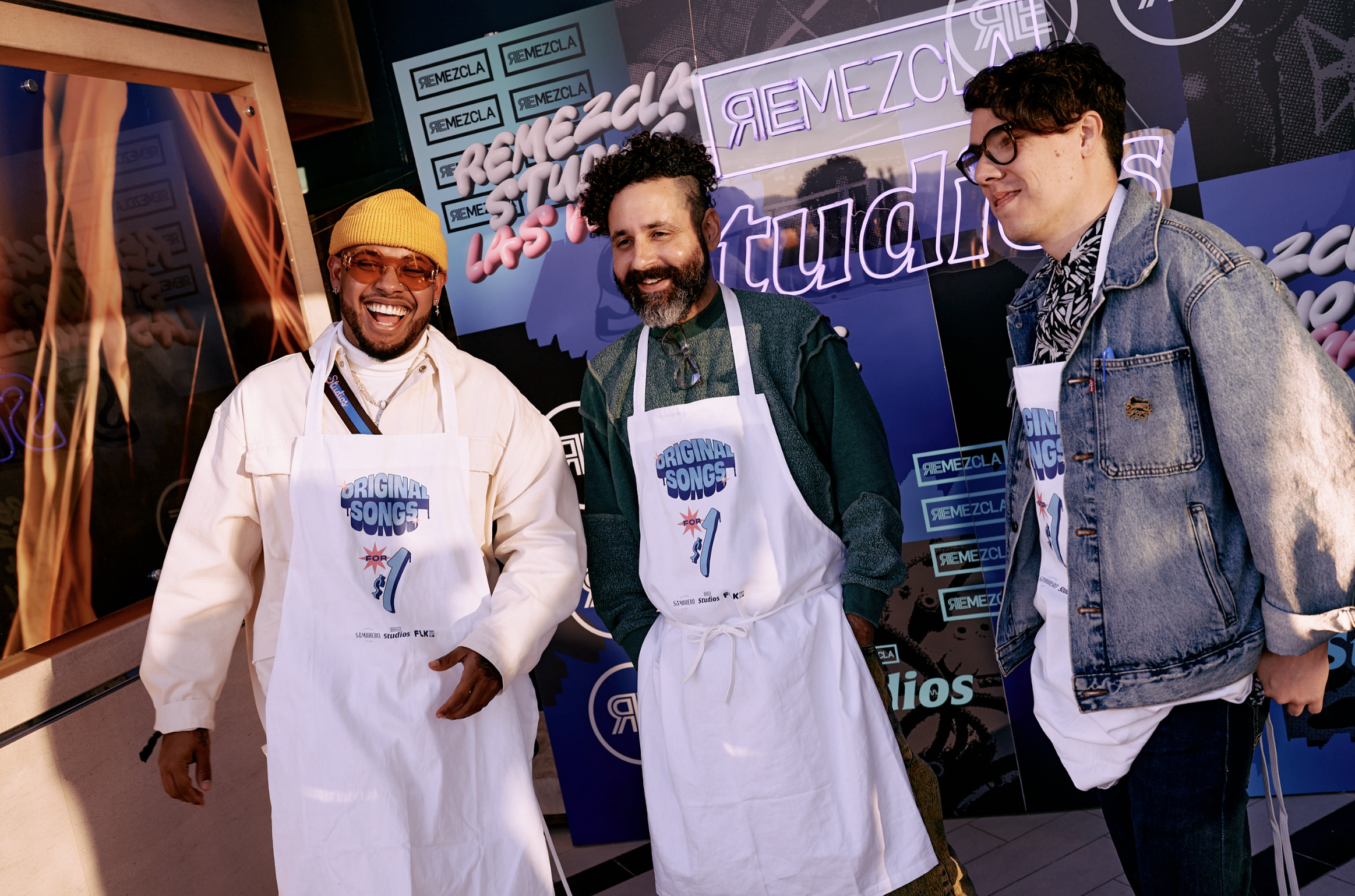 Tres hombres sonrientes en un evento, usan delantales blancos con el texto 'Original Songs for', uno de ellos lleva gafas y gorra amarilla, otro tiene barba y curly, y el tercero usa anteojos y una chamarra de mezclilla, frente a un fondo con logotip