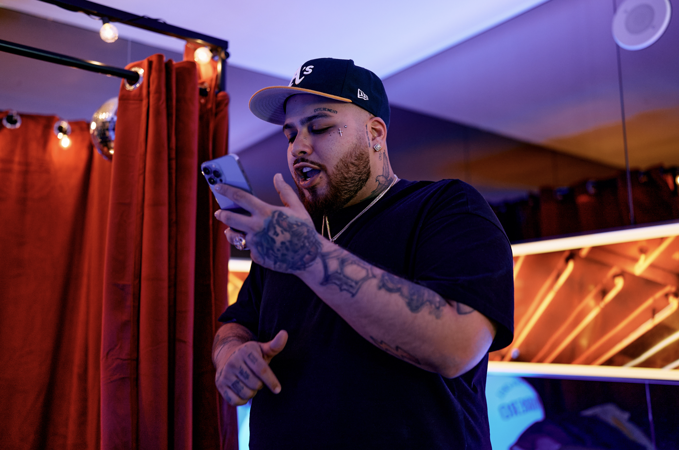 Hombre con tatuajes y barba, usando gorra y camiseta negra, mirando su teléfono en un ambiente con cortinas rojas y luces de neón.