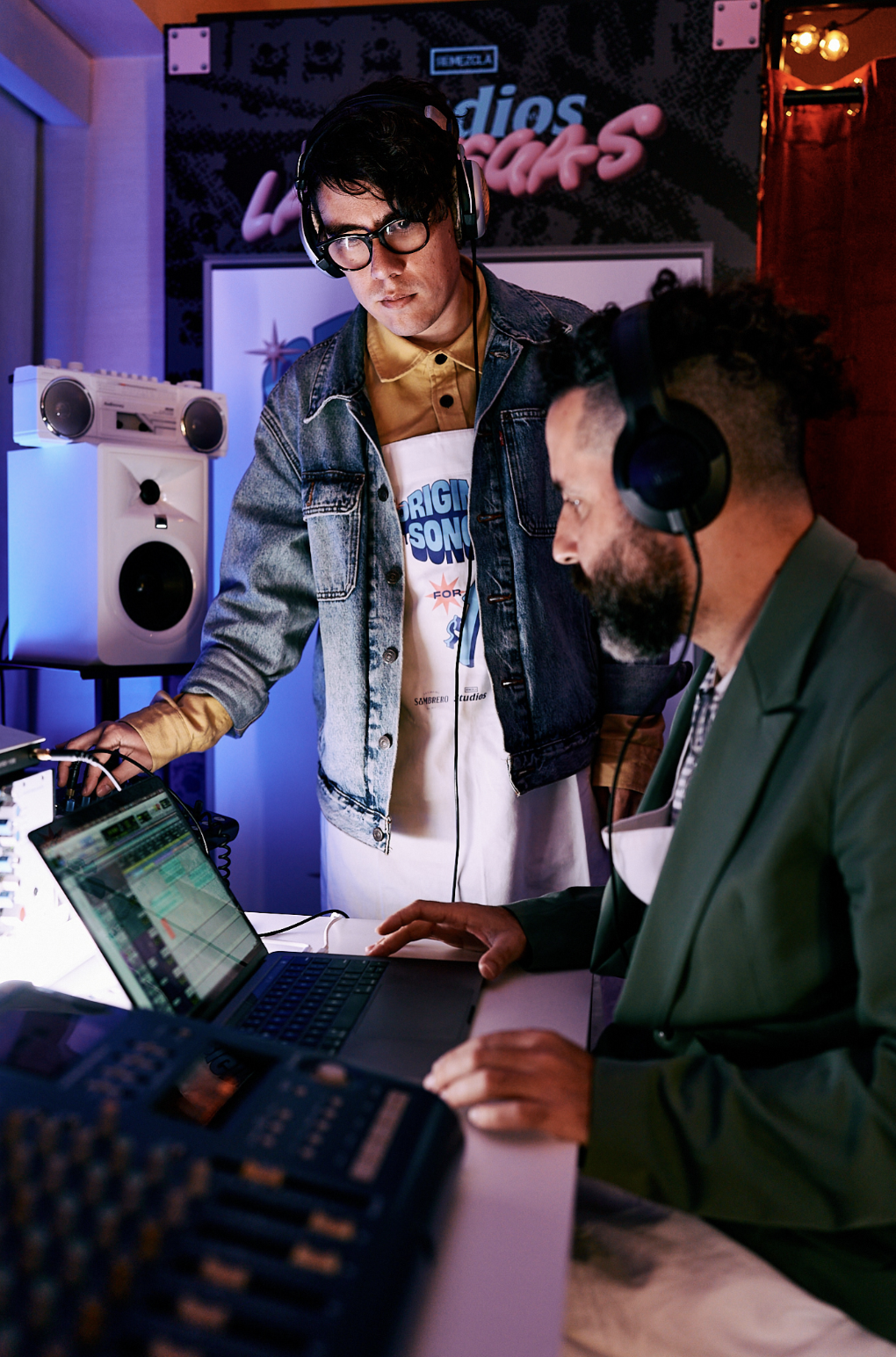 Dos hombres en un estudio de grabación, uno con gafas y chaqueta de mezclilla, el otro con barba y traje, trabajando con equipo de audio y computadora.
