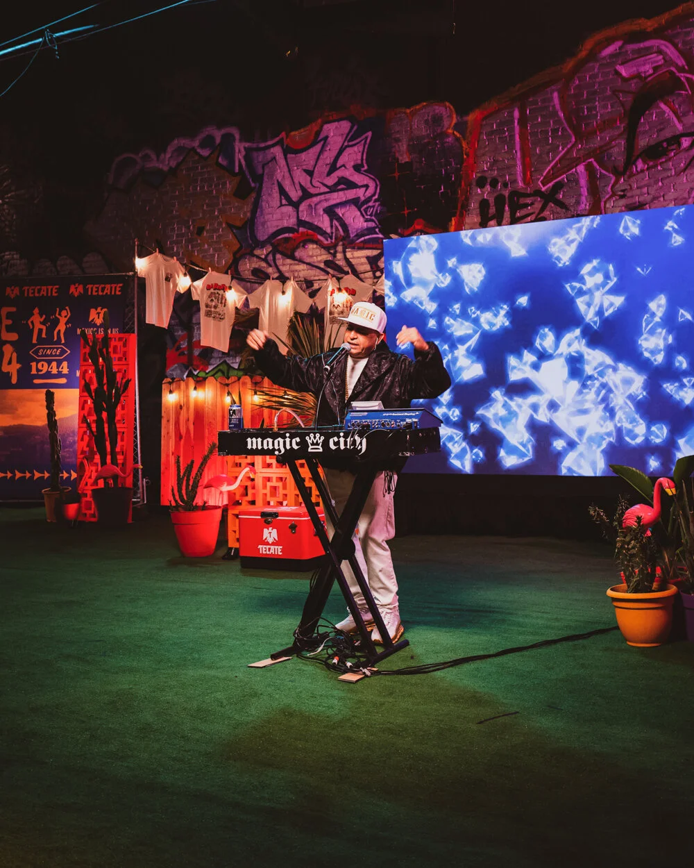 Un DJ tocando en un escenario decorado con cactuses, flamingos y arte urbano, con pantallas con luces y letras, en un ambiente festivo y colorido.