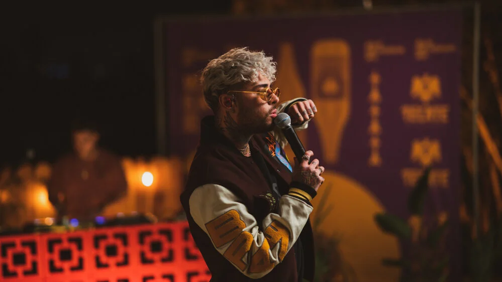 Un cantante con cabello gris, gafas naranjas, tatuajes y ropa oscura, sosteniendo un micrófono en un escenario de noche, con un DJ en el fondo y luces cálidas.