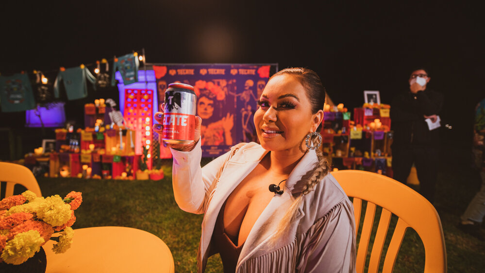 Mujer sonriendo y levantando una lata de cerveza en un evento con decoración colorida y personas en el fondo.