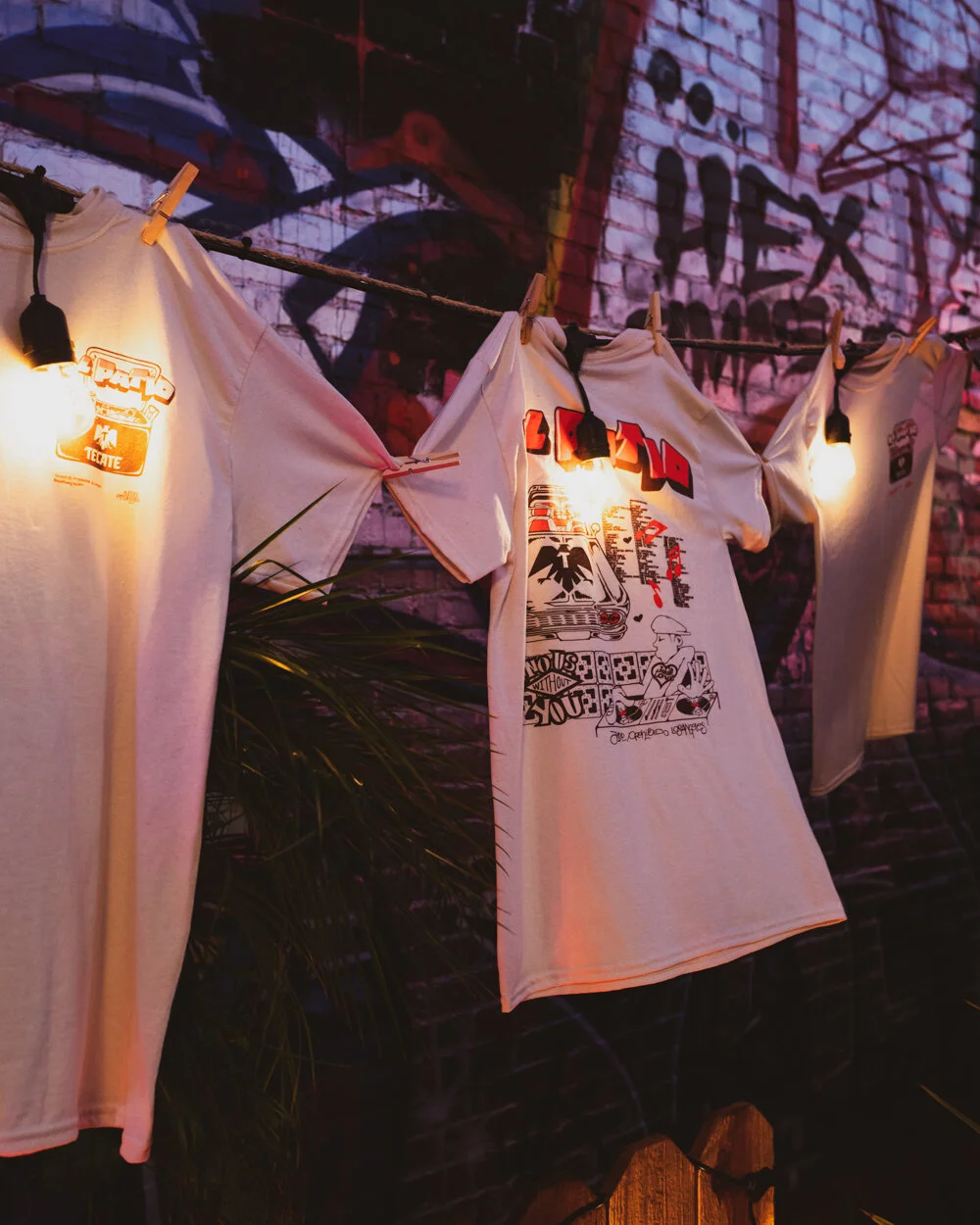 Camisetas blancas colgadas en una cuerda con pinzas, iluminadas por lámparas, con un mural de graffiti en la pared de fondo.