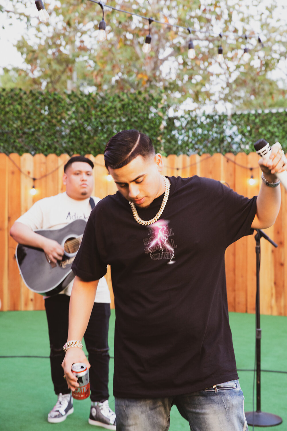 Jóvenes en un patio, uno con micrófono y otro tocando guitarra, decorado con luces y cerca de una cerca de madera, parece una fiesta o una reunión musical.