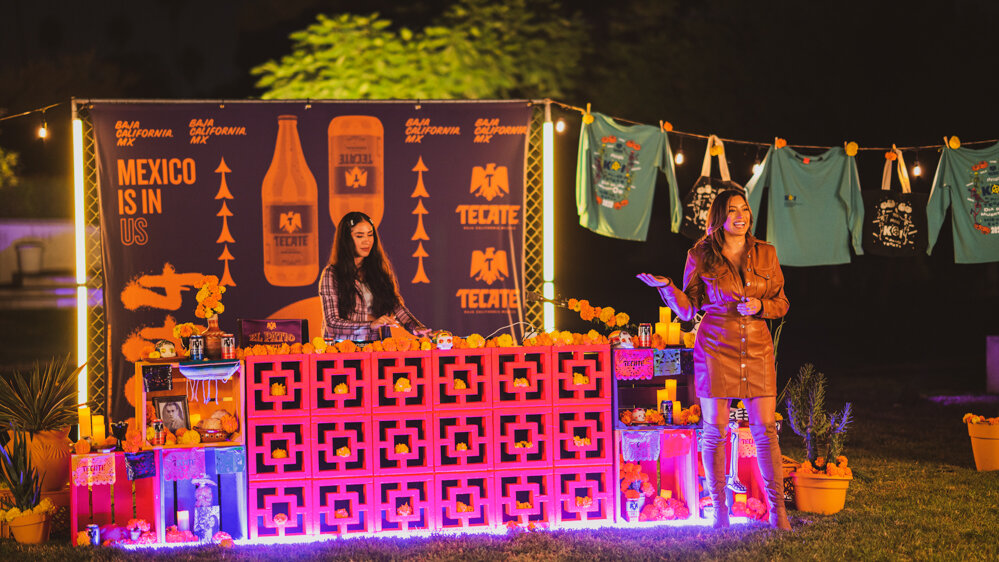 Una fiesta mexicana al aire libre con decoración colorida, una mesa con tequila y flores, y un banner que dice "México is in us" en fondo oscuro. Una mujer con abrigo de cuero y otra joven detrás de la barra.