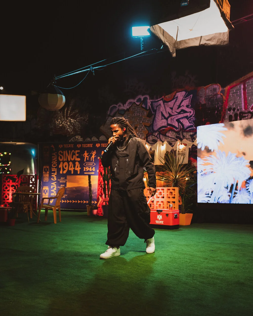 Una persona con ropa negra y zapatillas blancas canta con un micrófono en un escenario decorado con plantas, luces y graffiti en la pared, en un ambiente oscuro con iluminación vibrante.