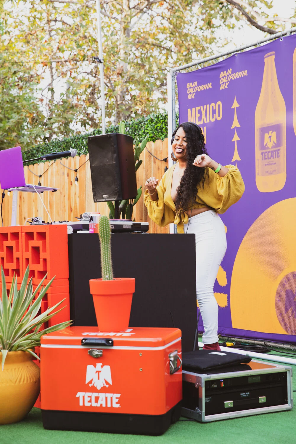 Mujer bailando en un evento al aire libre con decoración de colores y un cartel que dice 'México' y 'Tecate'.