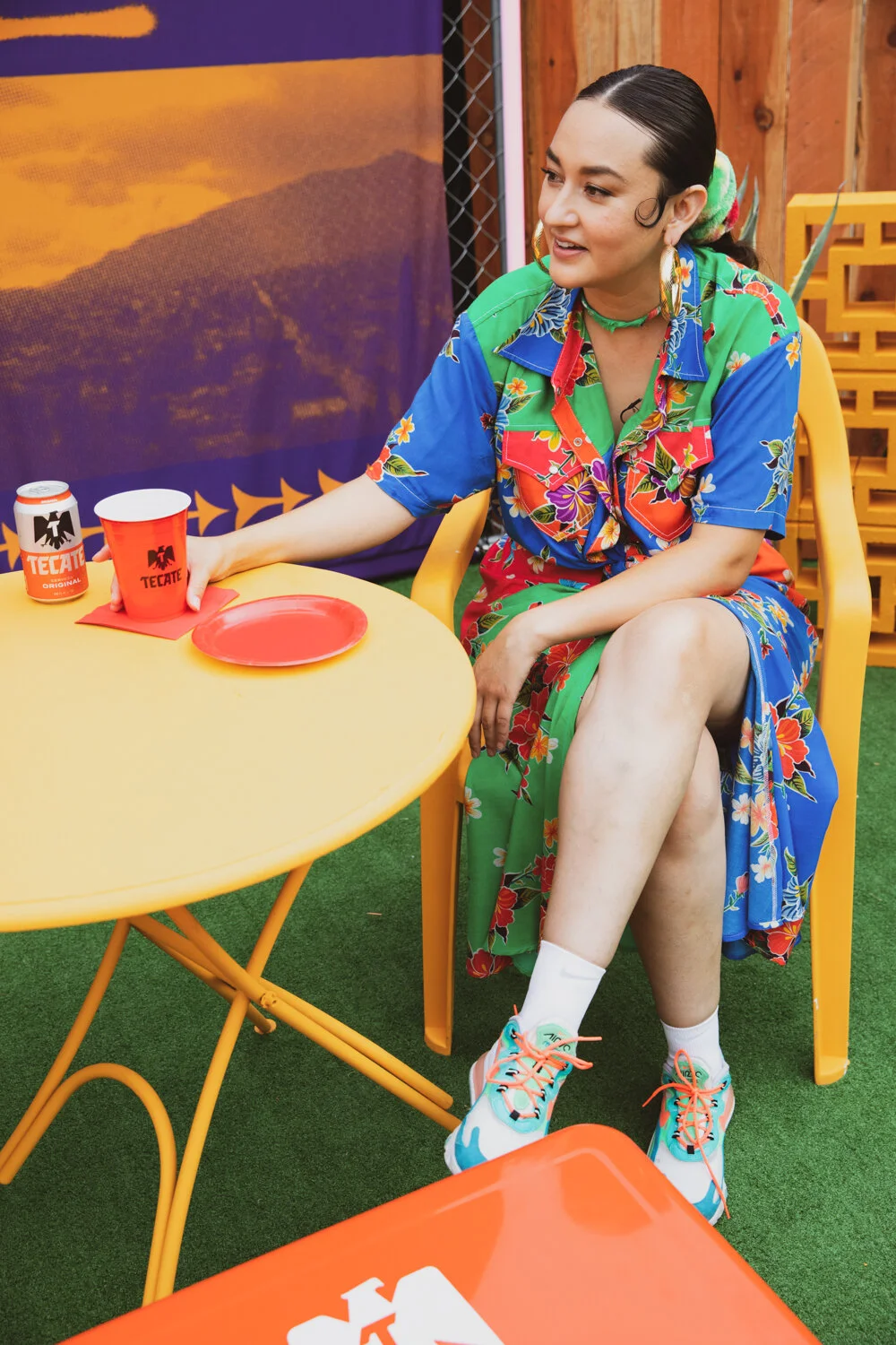 Mujer con cabello pulido, ropa colorida con flores, sentada en una silla amarilla en un espacio con decoración y muebles de madera, sosteniendo un vaso rojo en una mano y una lata de Tecate en la mesa.