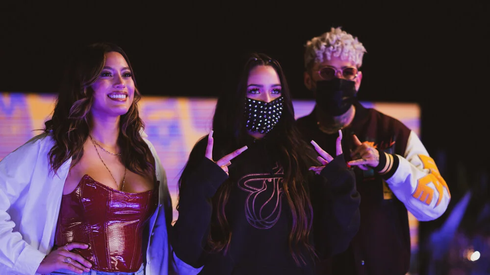 Tres personas en un evento nocturno, una mujer con vestido rojo y chaqueta blanca, una mujer con mascarilla de perlas y un hombre con gafas rosadas y chaqueta moderna.